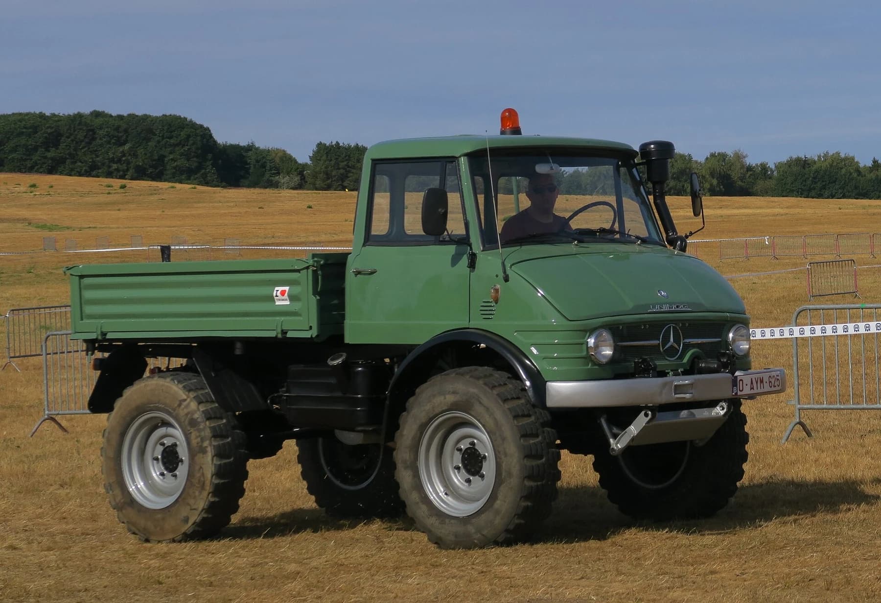 A restored green Unimog 406 with a short cab, open load bed and large tyres, seen from the front quarter on a dry field.