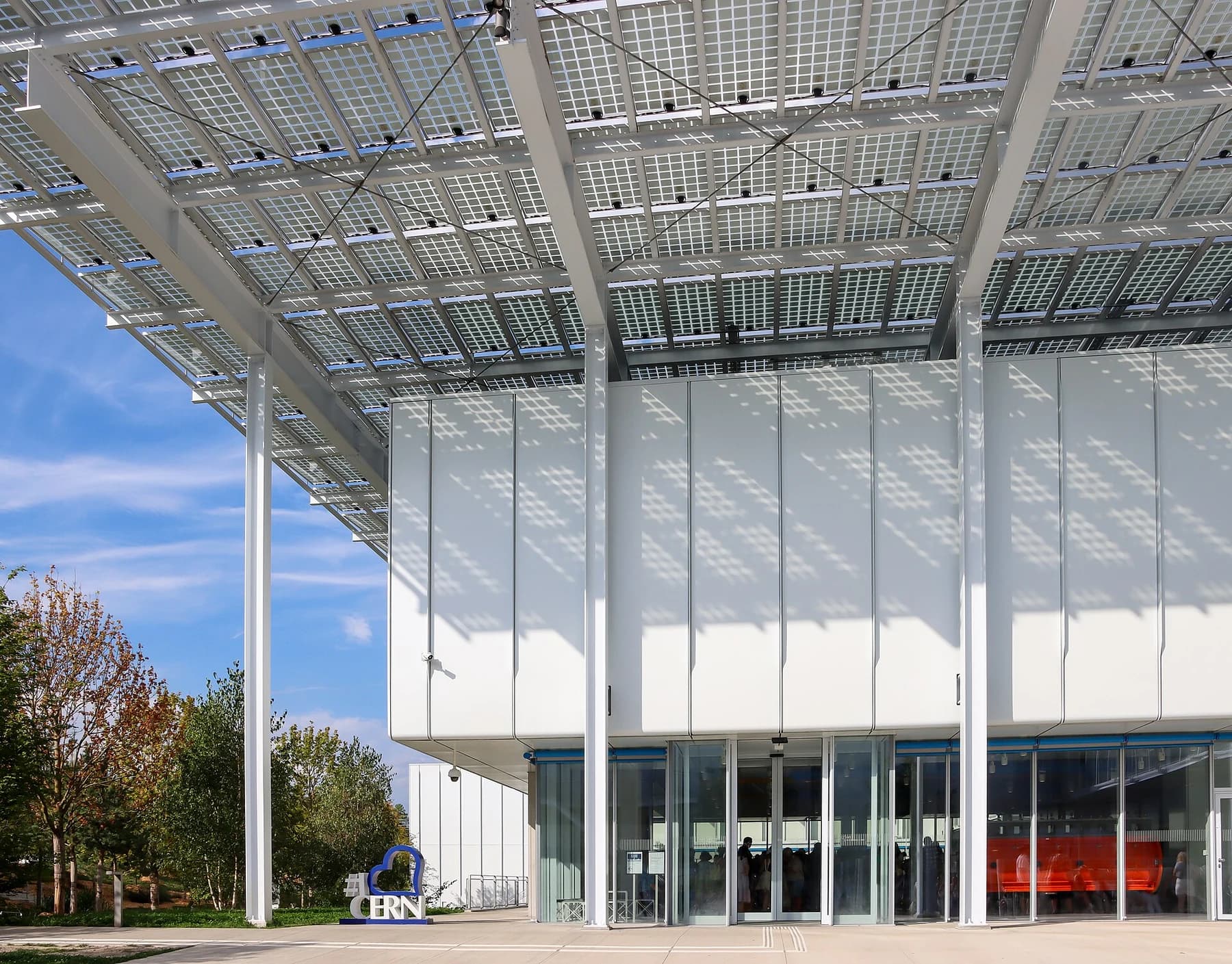CERN Science Gateway’s glass-fronted public entrance beneath an elevated solar-panel roof, photographed in daylight with trees beside the building.
