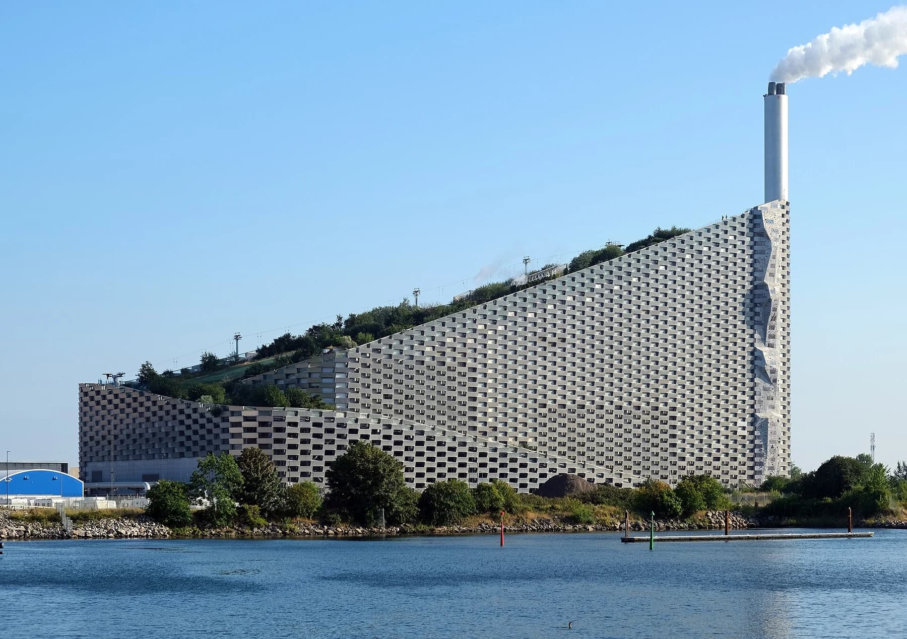 CopenHill and its tall chimney beside the water in Copenhagen, with the plant’s sloping planted roof and patterned metal façade visible in daylight.