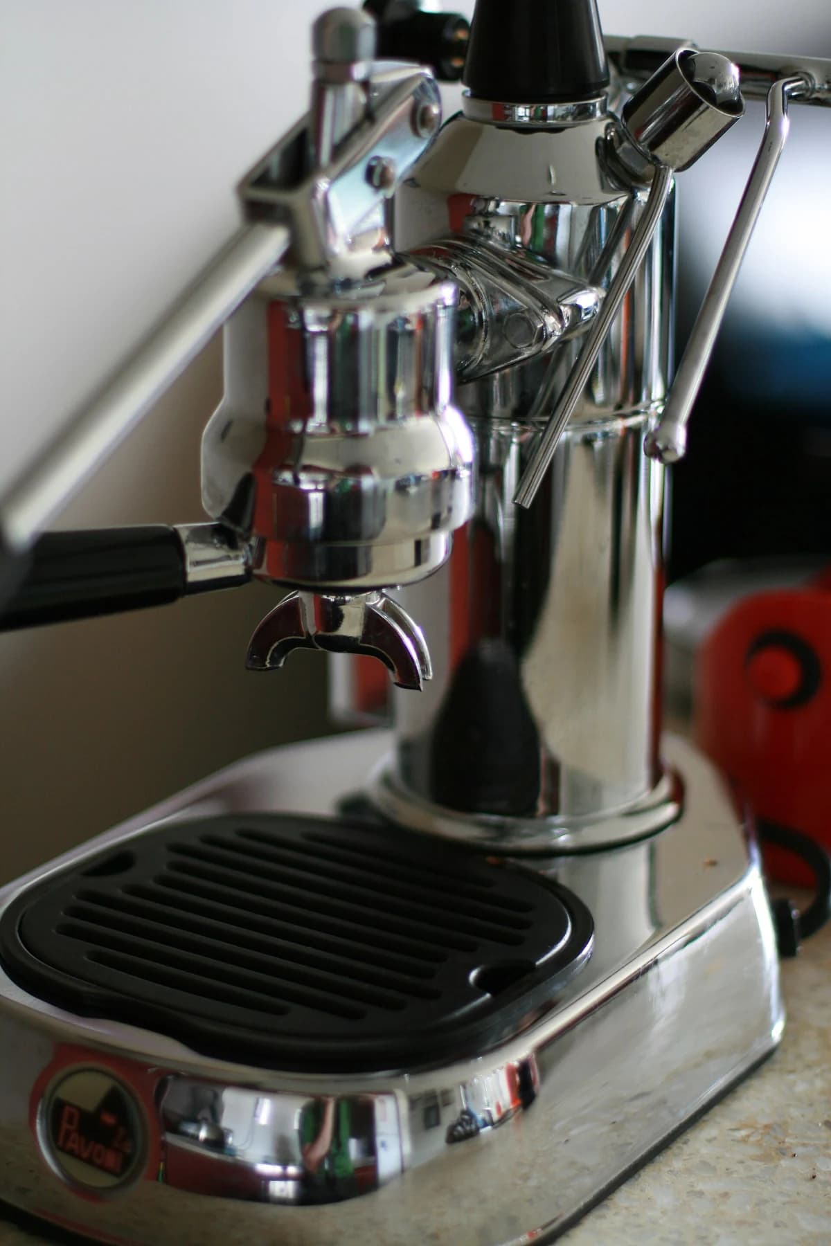 A close detail of a chrome La Pavoni Europiccola, showing its brewing group, black-handled portafilter and drip tray in a domestic setting.