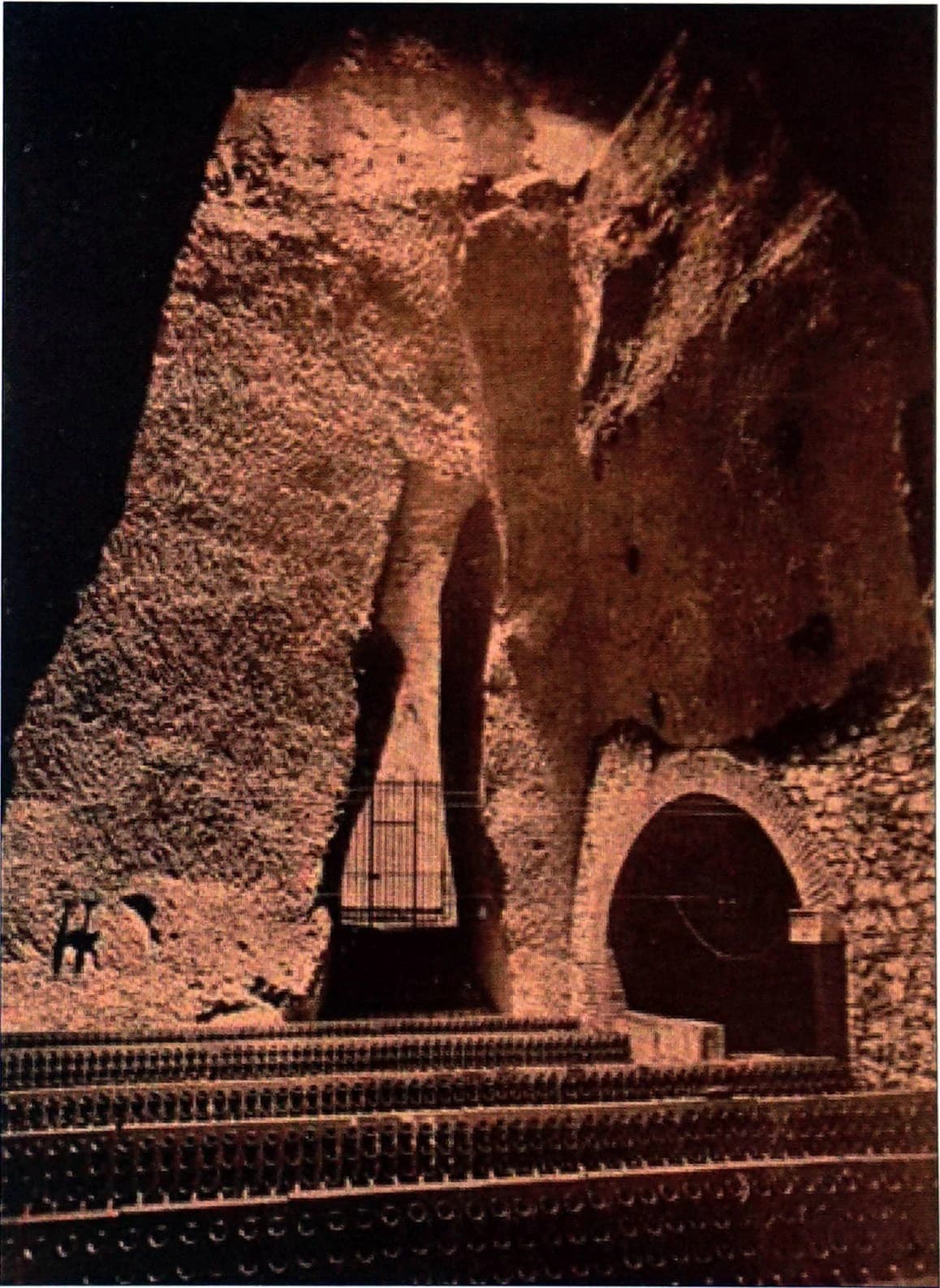 An archival view inside Ruinart’s chalk cellars in Reims, with warmly lit rough stone, a high opening and rows of bottles near the floor.