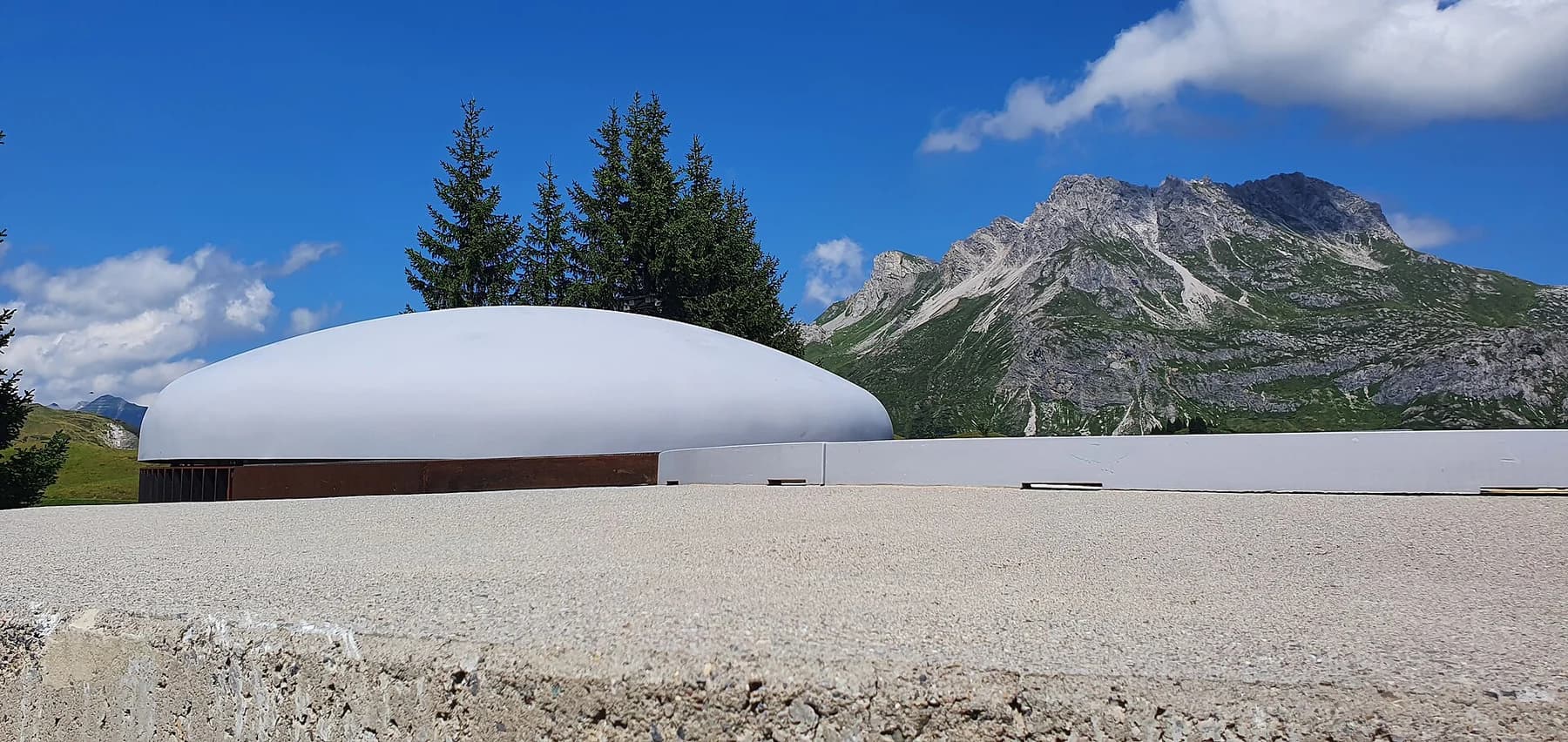 The pale dome of James Turrell’s Skyspace Lech seen above a low wall, with conifers, blue sky and an Austrian mountain rising behind it.