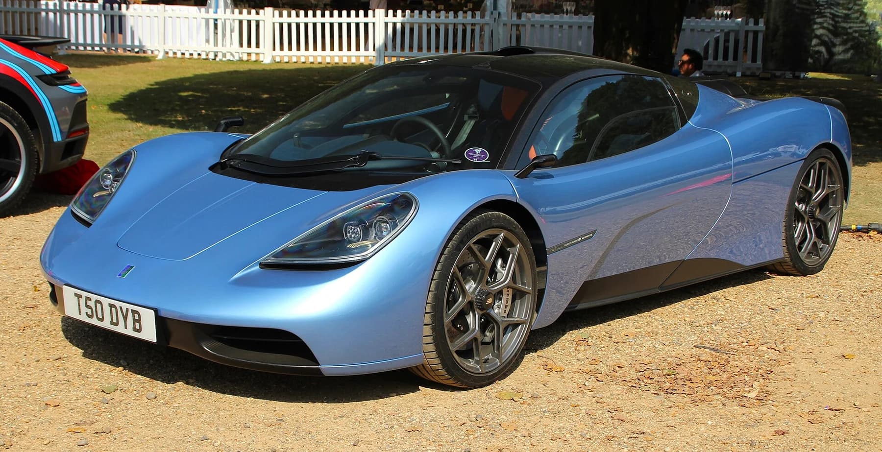 A blue road-going Gordon Murray Automotive T.50 at Hampton Court, seen from the front quarter with its compact body and central cabin visible.