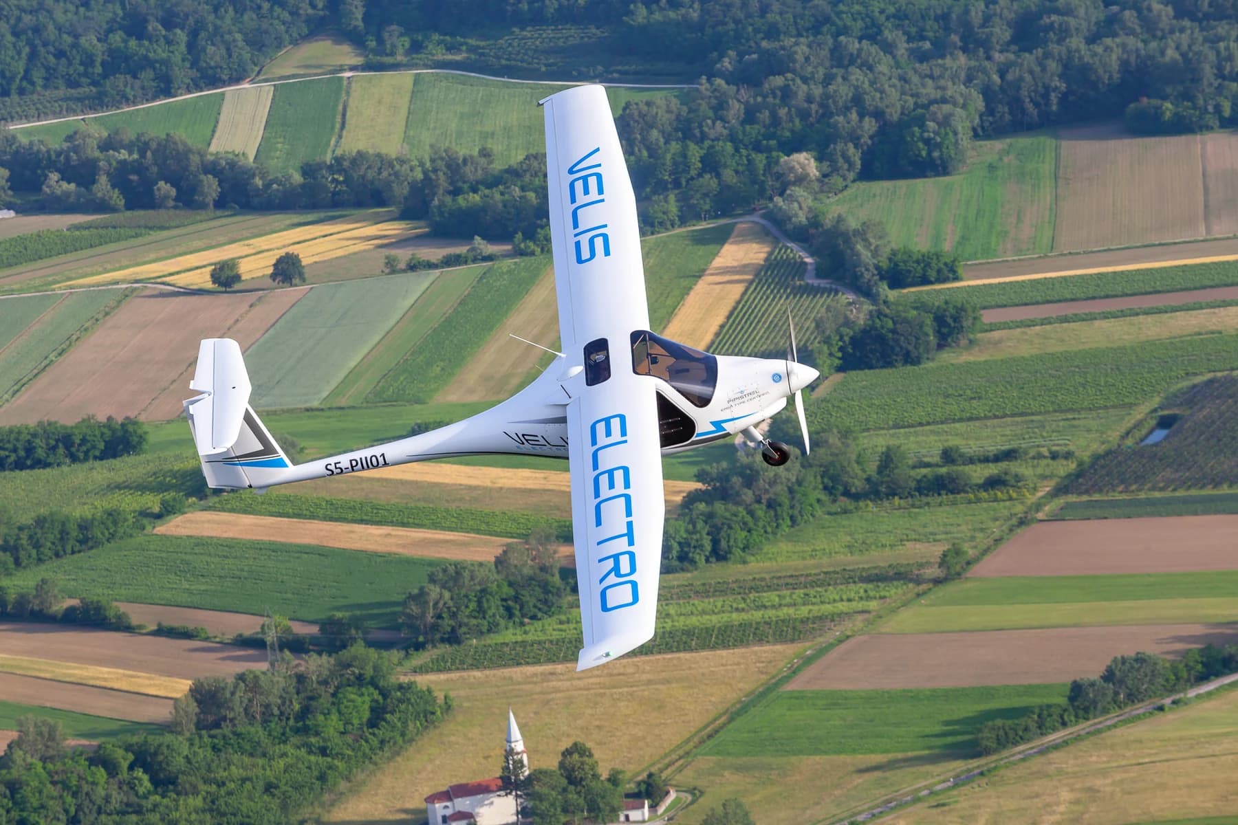 A white Pipistrel Velis Electro banking in flight above green fields, with blue markings and Velis Electro lettering across its wings.