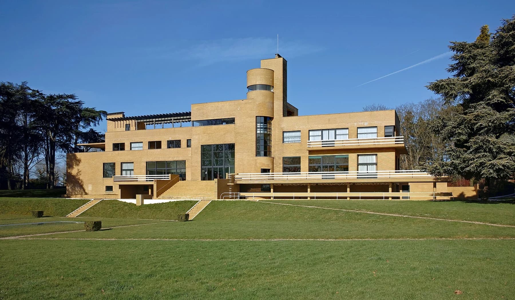 Villa Cavrois seen across its green garden, with long yellow-brick wings, horizontal windows and a rounded tower against a clear blue sky.