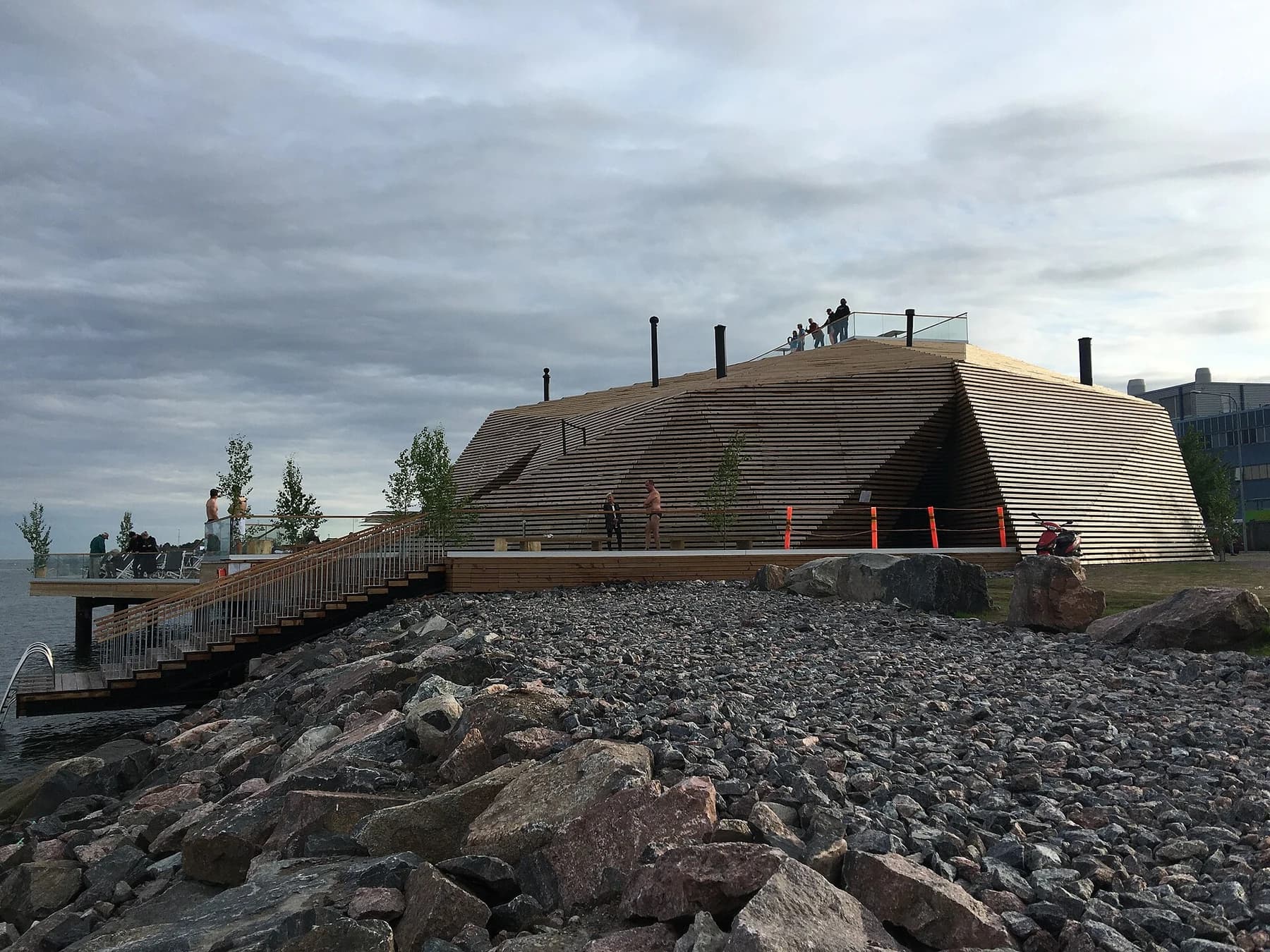 Löyly’s angular timber sauna building and roof terrace beside Helsinki’s rocky waterfront beneath a cloudy sky.