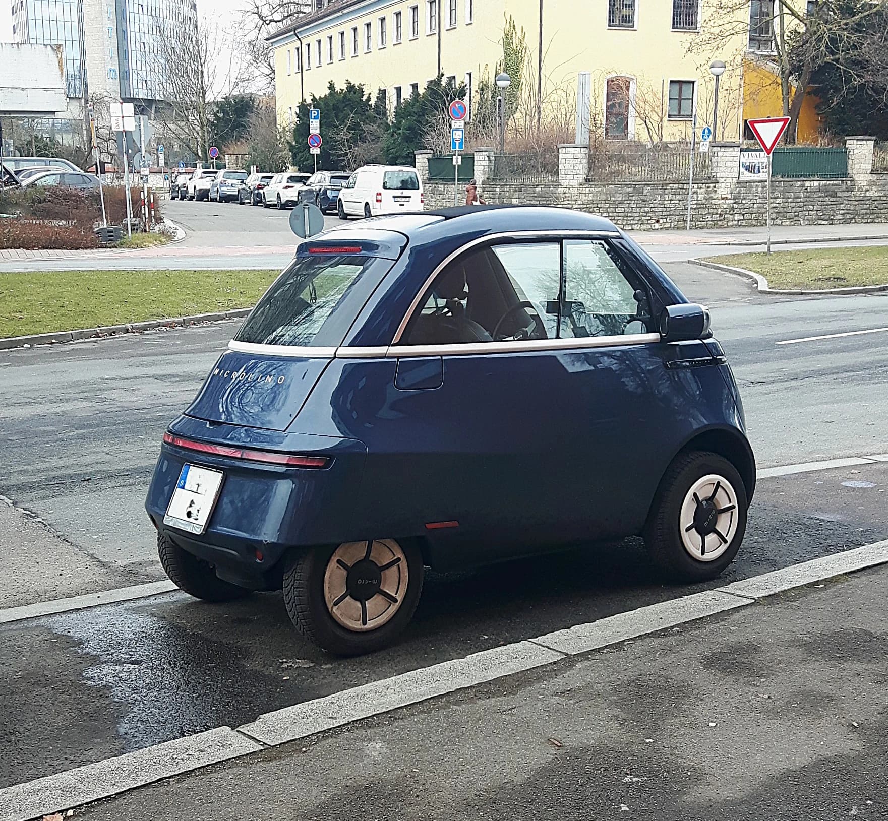 A dark blue Microlino parked beside a street in Neu-Ulm, showing its compact enclosed body and small wheels.