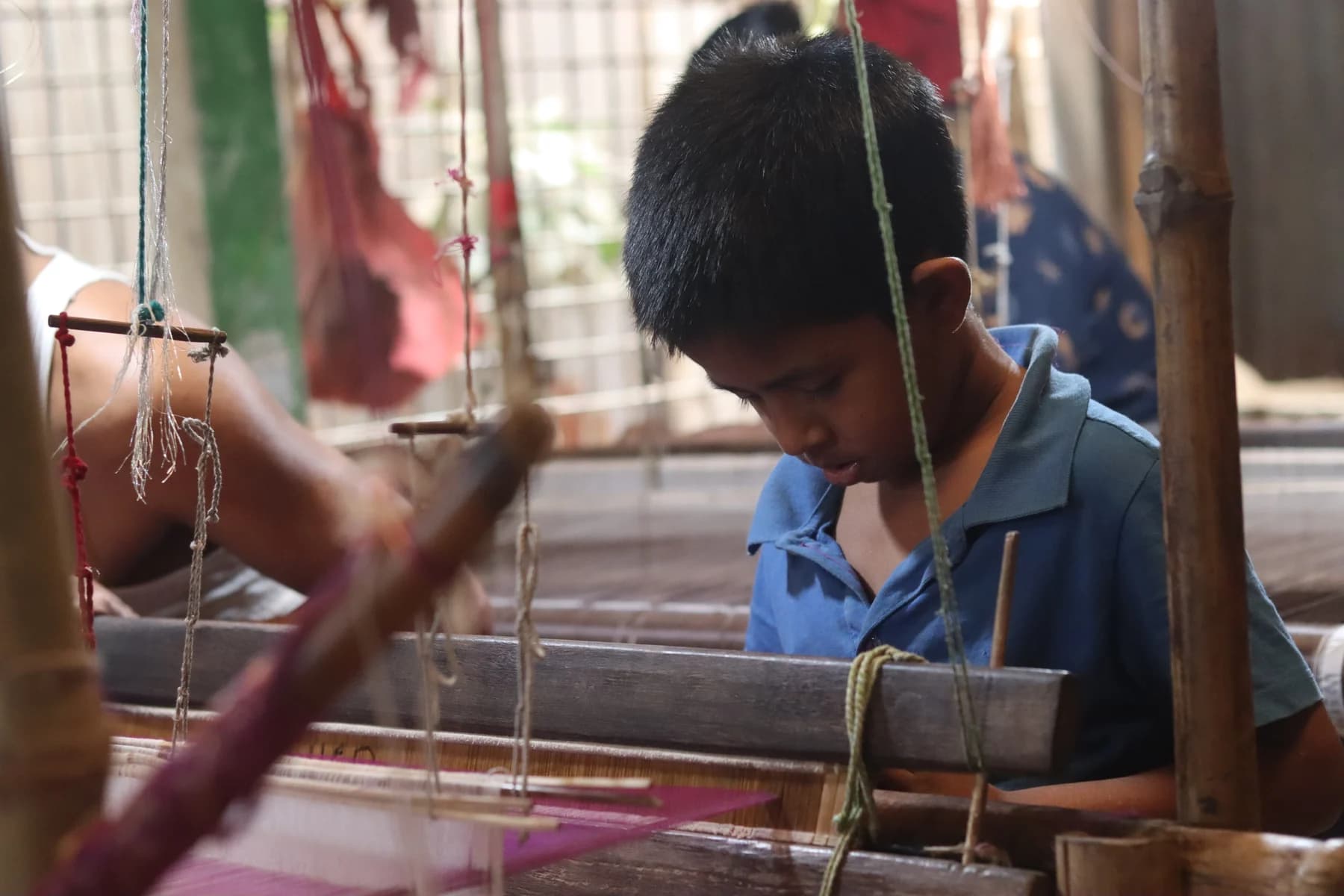 A Jamdani weaver concentrates on the threads of a loom in a workshop, with another worker’s hand visible in the foreground.