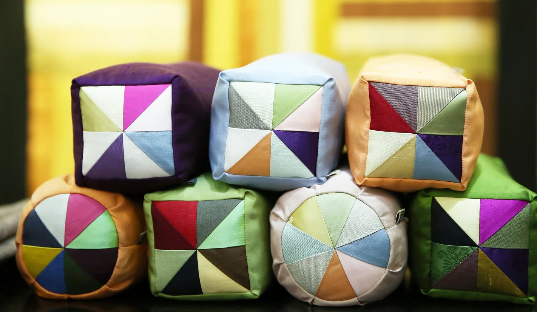 Small textile pillows display pinwheel arrangements of triangular fabric pieces in green, violet, pale blue, red and gold.