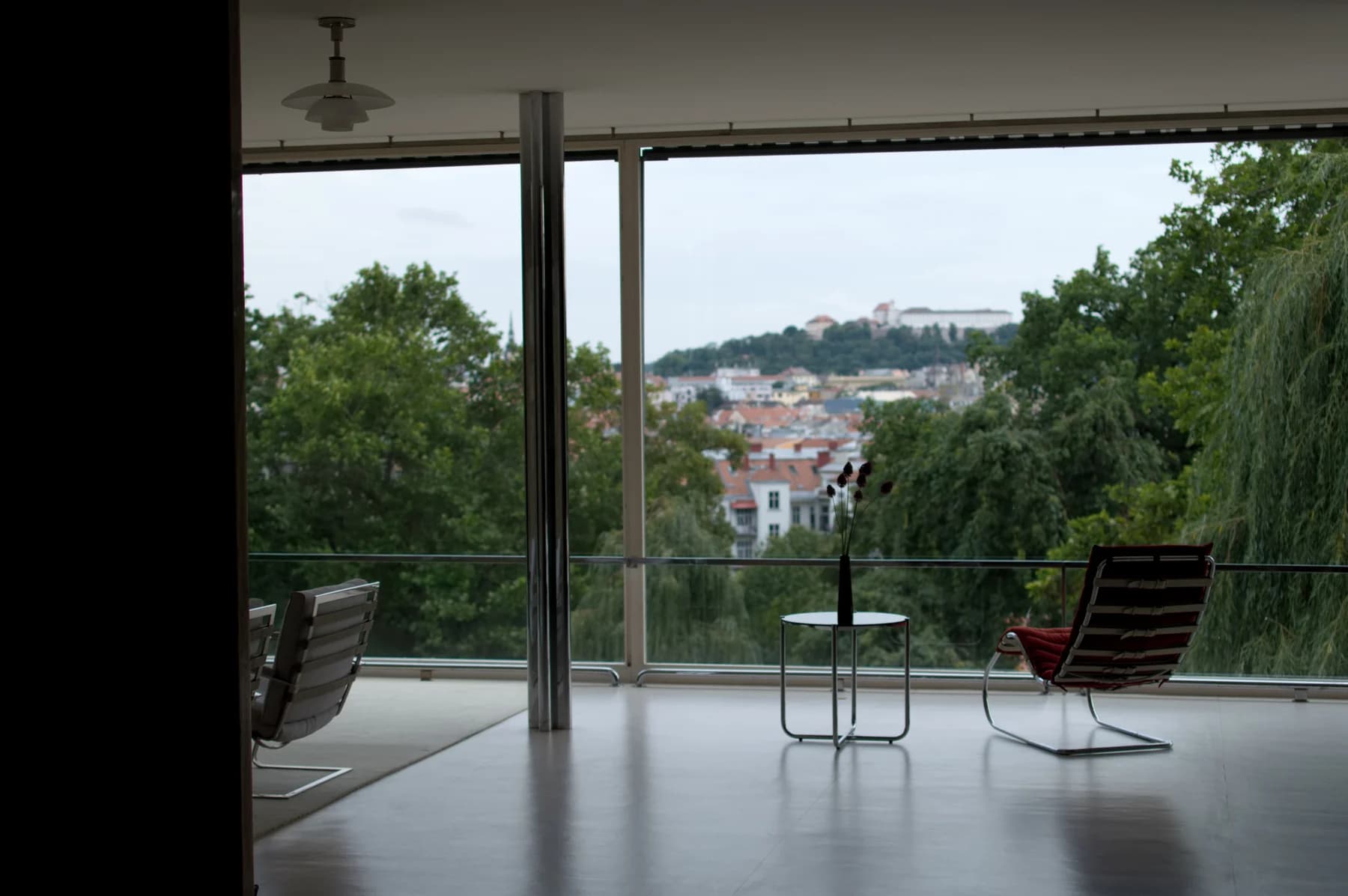Villa Tugendhat’s living area opens through broad glazing towards the garden and Brno, with chairs and a small table in the foreground.