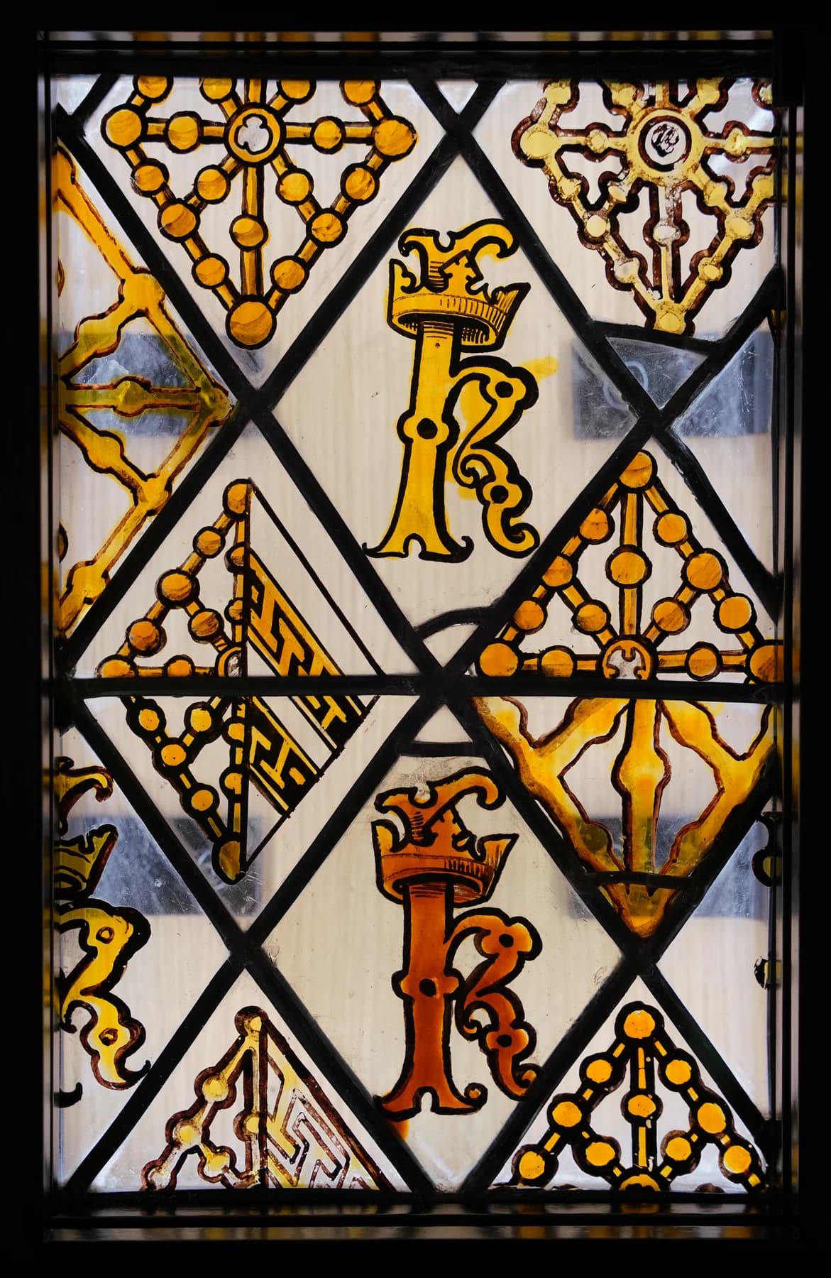 A stained-glass panel combines crowned letter forms, gold ornaments and pale diamond-shaped pieces held together by dark joints.