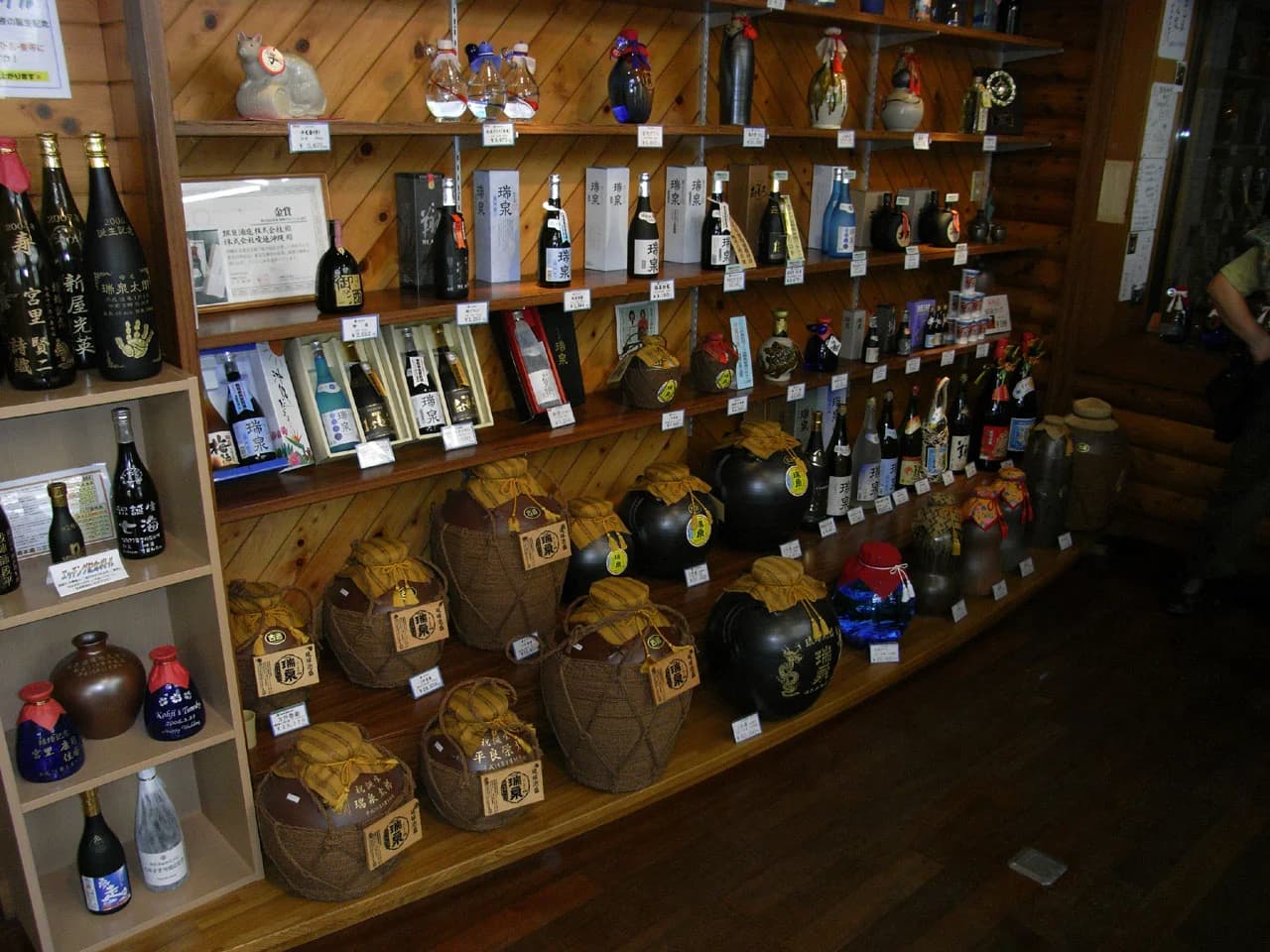 Zuisen’s showroom shelves hold dark ceramic awamori vessels, some wrapped in woven coverings, alongside boxed bottles.