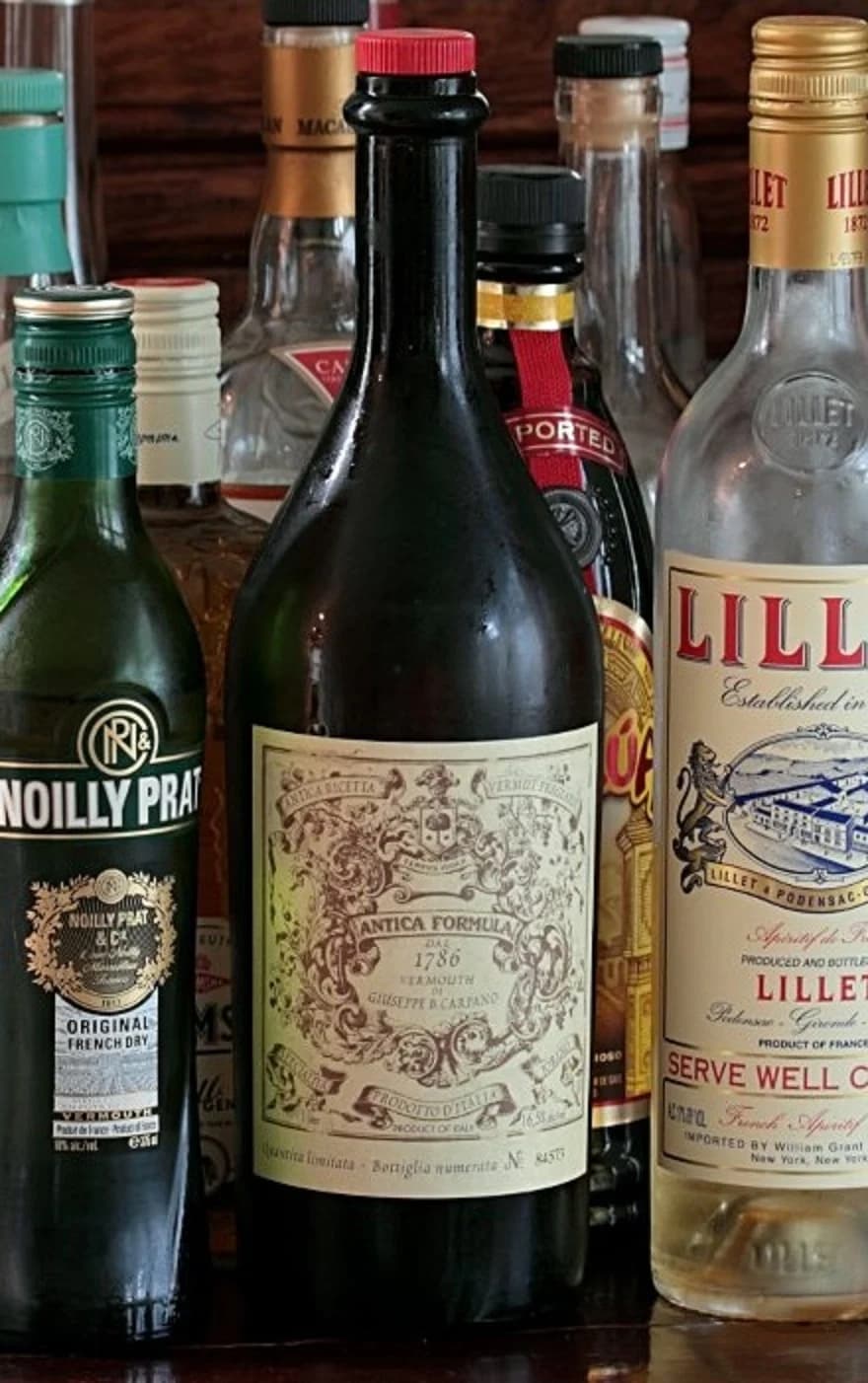 A dark Carpano Antica Formula bottle with an ornate cream label and red cap standing among other vermouth and aperitif bottles.