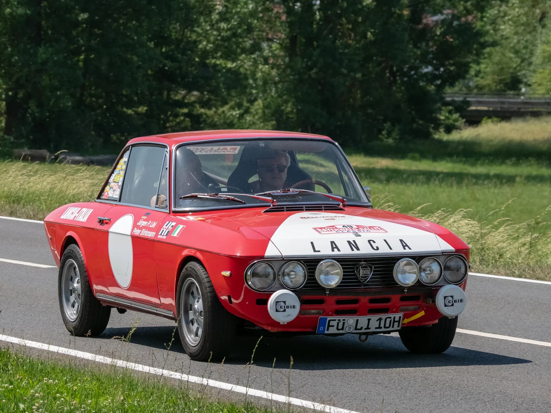 A red Lancia Fulvia coupé with white rally-style panels and additional front lamps driving past grass and trees.