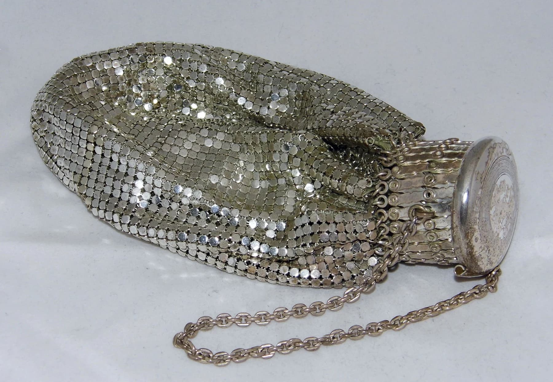 A silver-coloured Whiting & Davis mesh purse lying on a pale surface, with a rounded rigid top and a fine chain beside it.