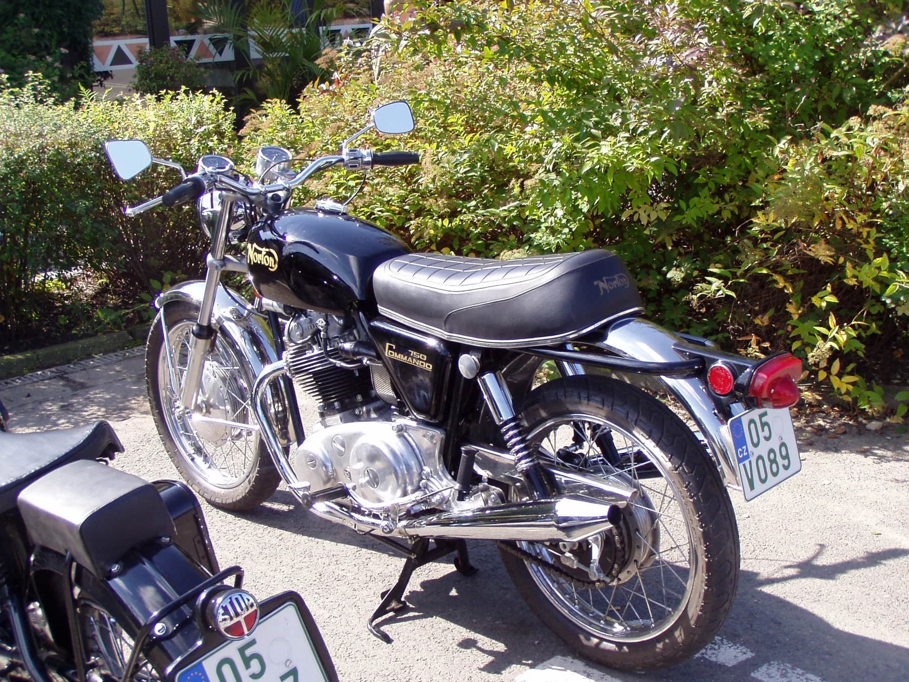 A black Norton Commando 750 seen from the rear left beside garden shrubs, with its twin-cylinder engine, chrome exhaust and spoked wheels clearly visible.