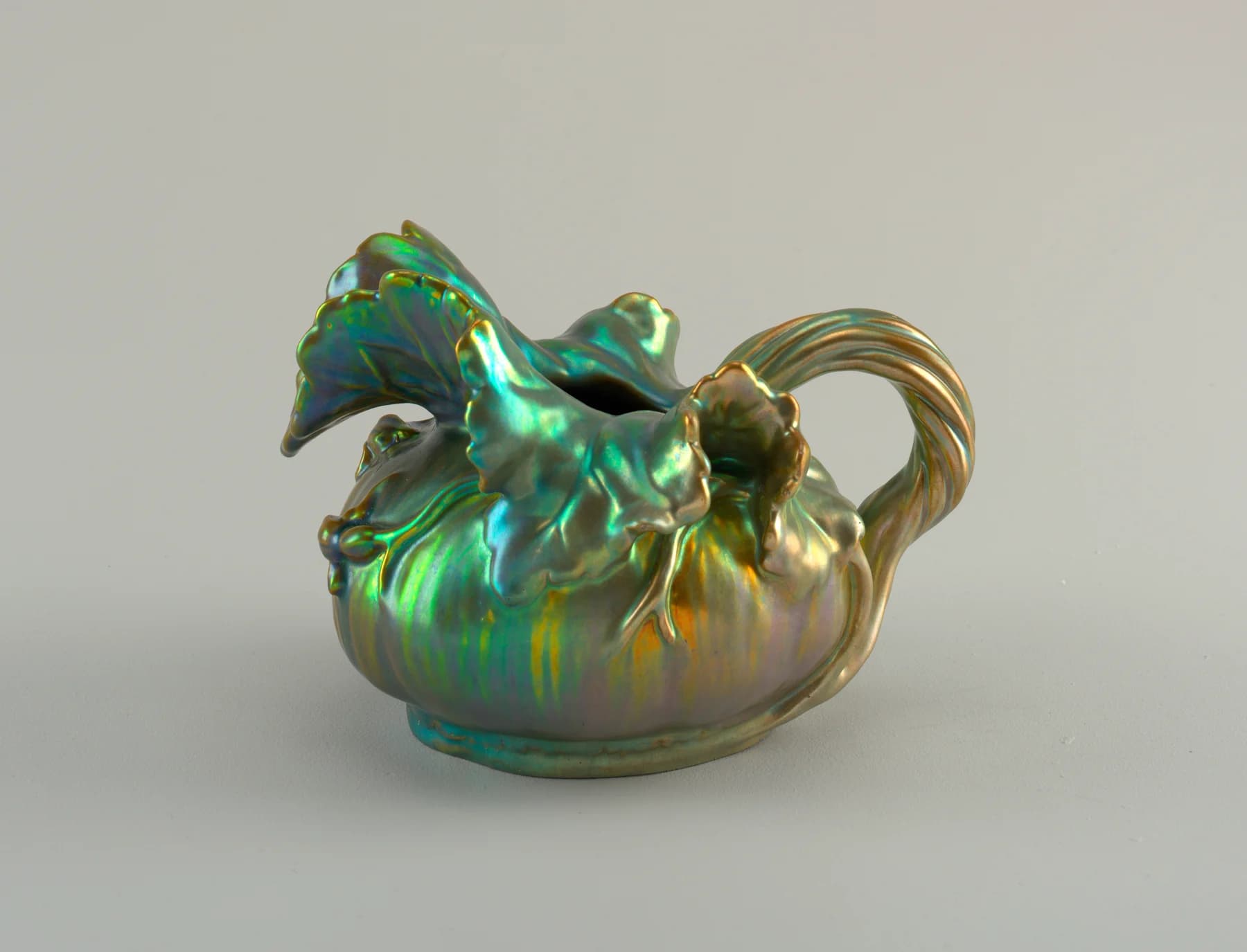 A rounded Zsolnay ceramic pitcher with a large leaf-shaped spout, a ribbed handle and an iridescent green-and-gold surface.