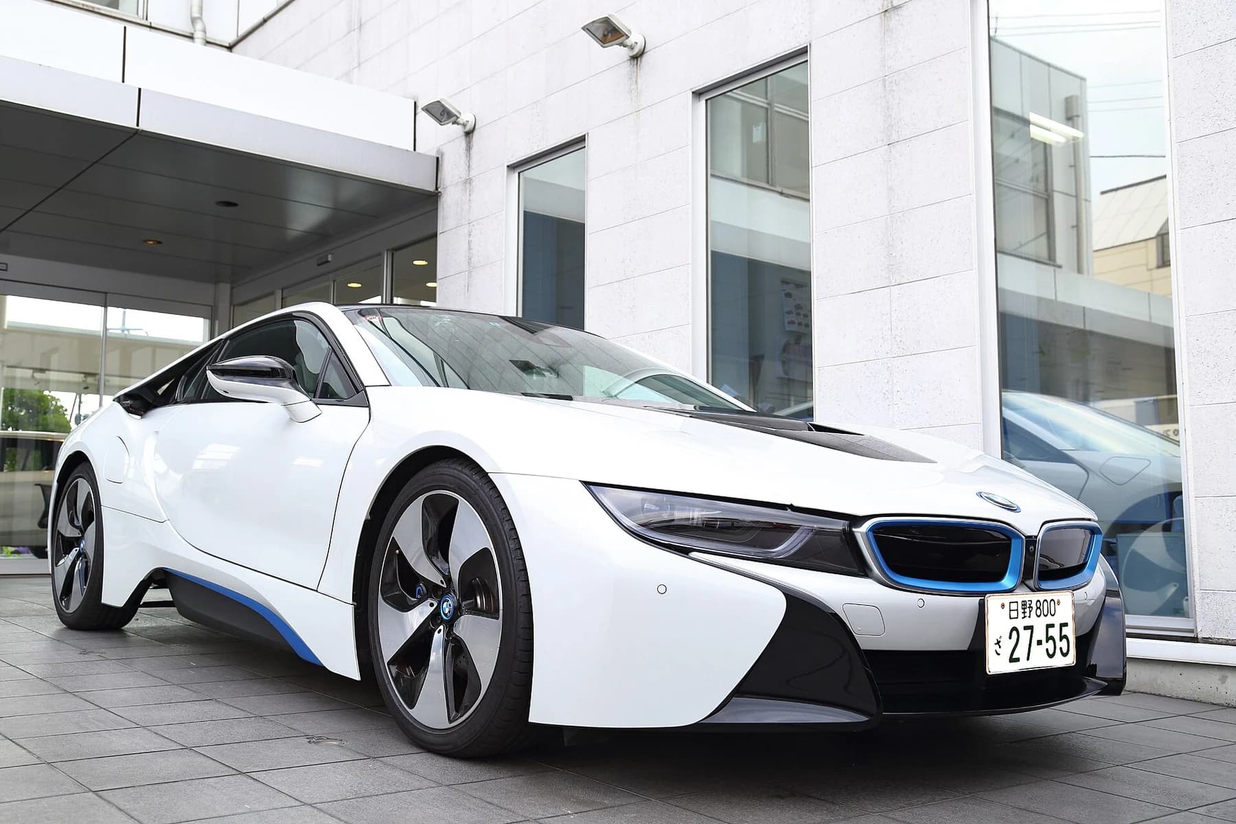 A white BMW i8 production coupé viewed from the front left outside a dealership, showing its low body, dark bonnet and blue lower-body accents.