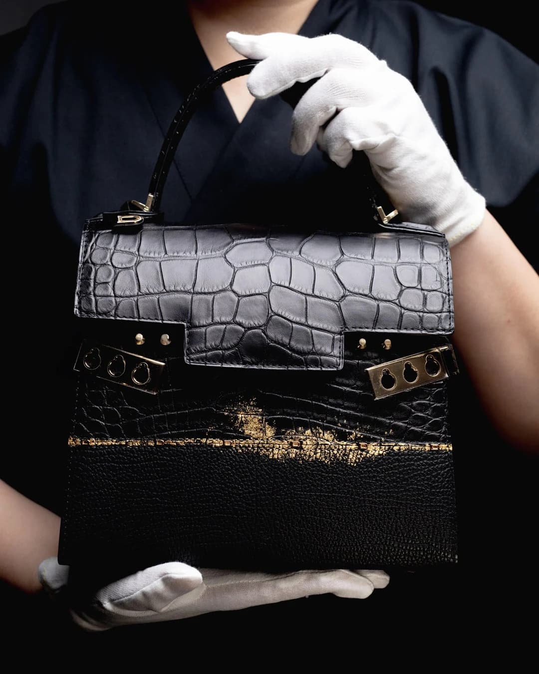 A black Delvaux Tempête bag held in white-gloved hands, with gold leaf tracing a horizontal join below its distinctive metal fittings.