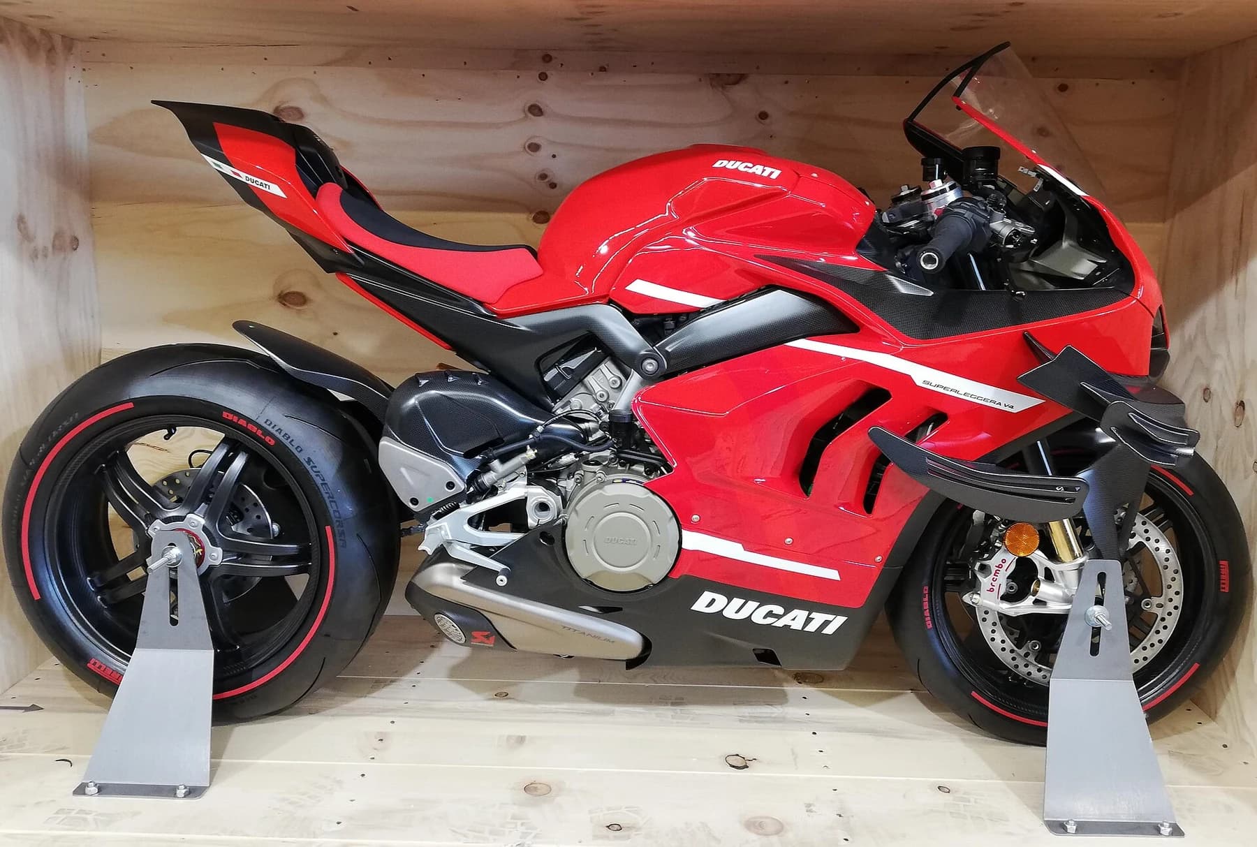 A red and black Ducati Superleggera V4 viewed from the side in its opened wooden transport crate, with carbon wheels and sculpted bodywork visible.
