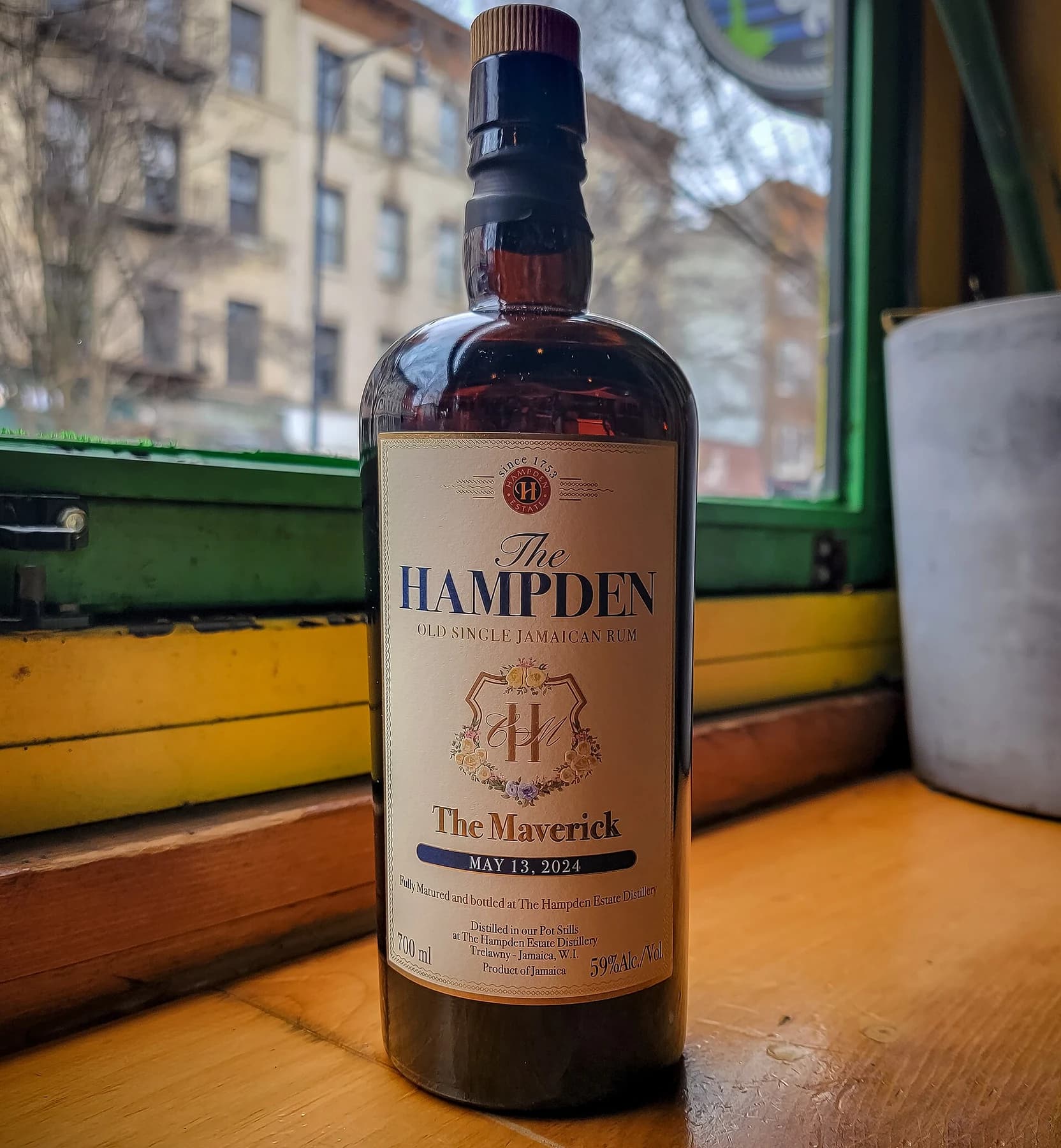 A bottle of Hampden Estate The Maverick on a wooden table beside a green window, with its cream label and commemorative date visible.