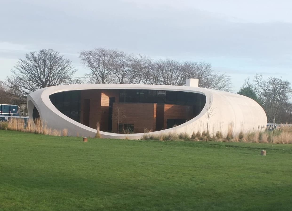 Maggie’s Aberdeen seen across a green lawn, with its low white curved shell framing a broad dark window and warm timber interior.