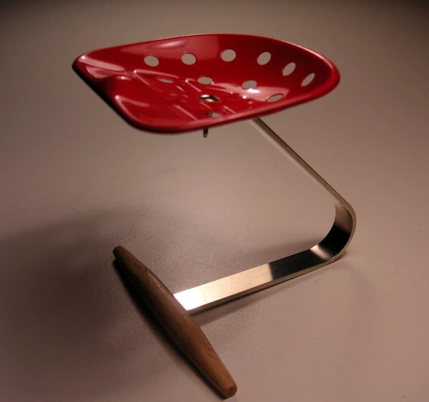 A red Mezzadro stool showing its perforated metal tractor-seat form, curved chrome support and wooden crosspiece against a warm neutral background.