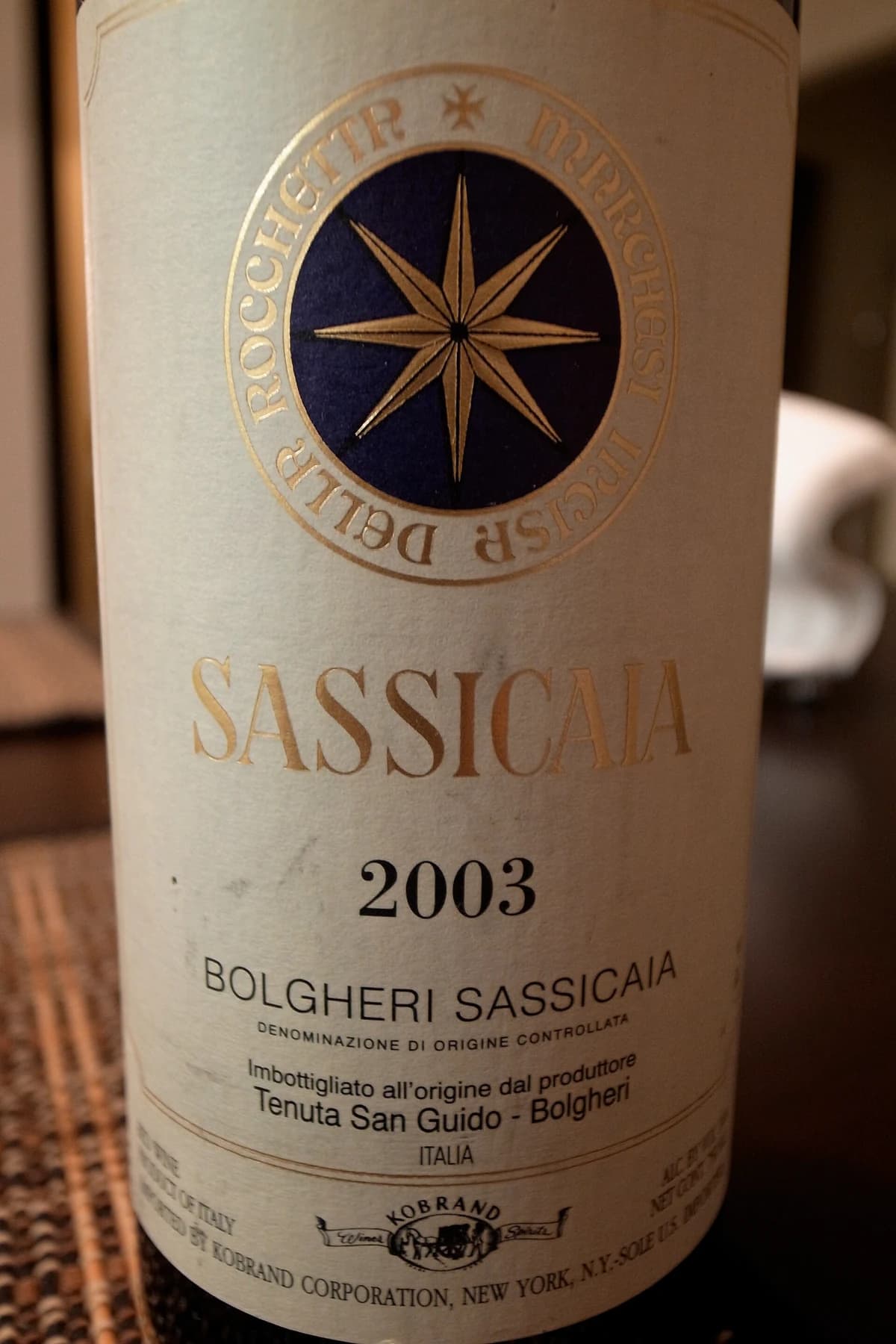 A close view of a Sassicaia 2003 wine label, with its gold compass-like star, vintage and Bolgheri Sassicaia denomination in warm light.