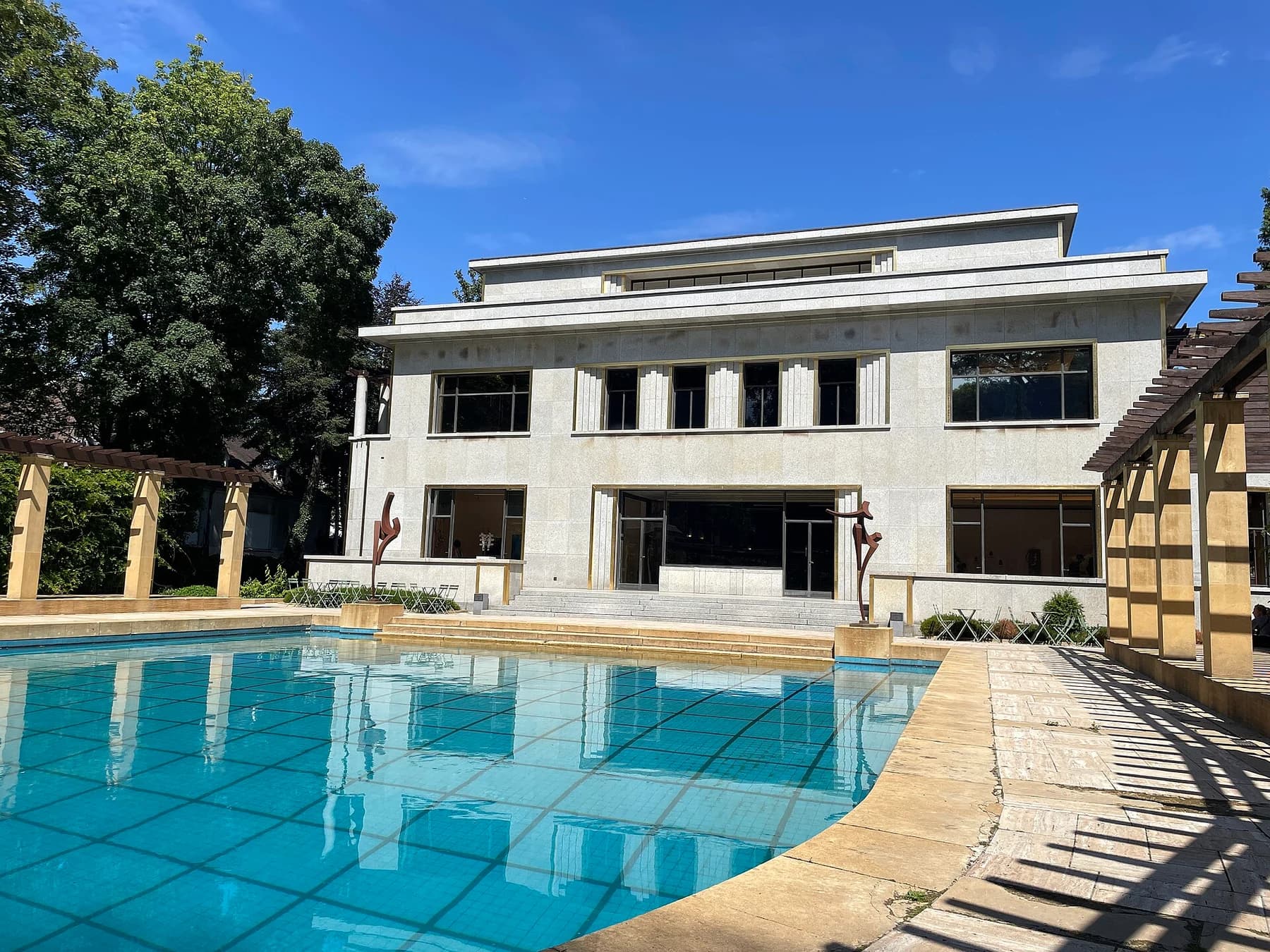 The rear facade and blue swimming pool of Villa Empain in Brussels, with pale stone terraces and dark-framed windows under a summer sky.