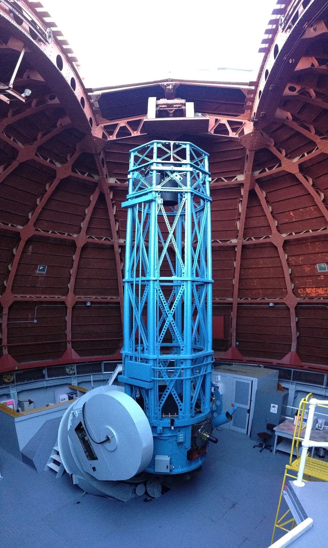 Mount Wilson’s blue 60-inch telescope extending vertically beneath the open slit of its brown observatory dome, with the instrument’s support machinery below.