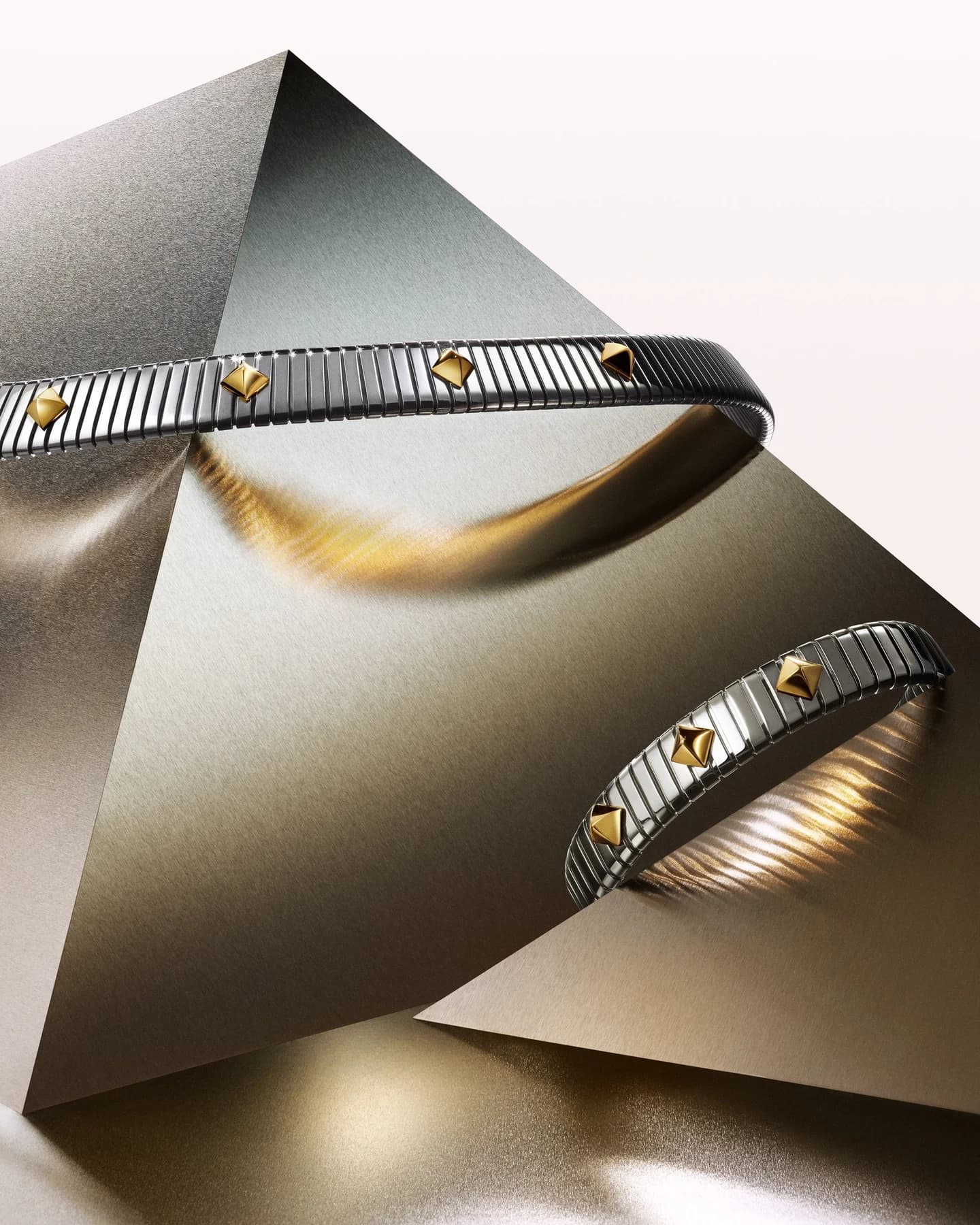 Bulgari Tubogas Gold & Steel necklace and bracelet arranged on a reflective metallic surface, with yellow-gold accents interrupting the steel coils.