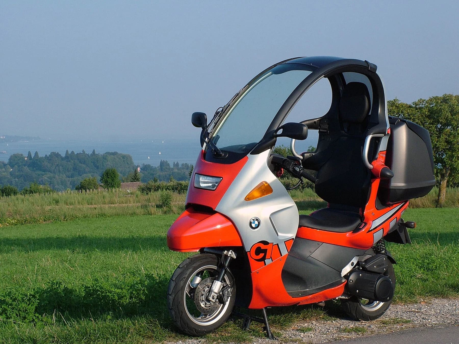 A red and silver BMW C1 parked beside a green field, showing its roof, windscreen, tall seat and narrow two-wheeled layout.