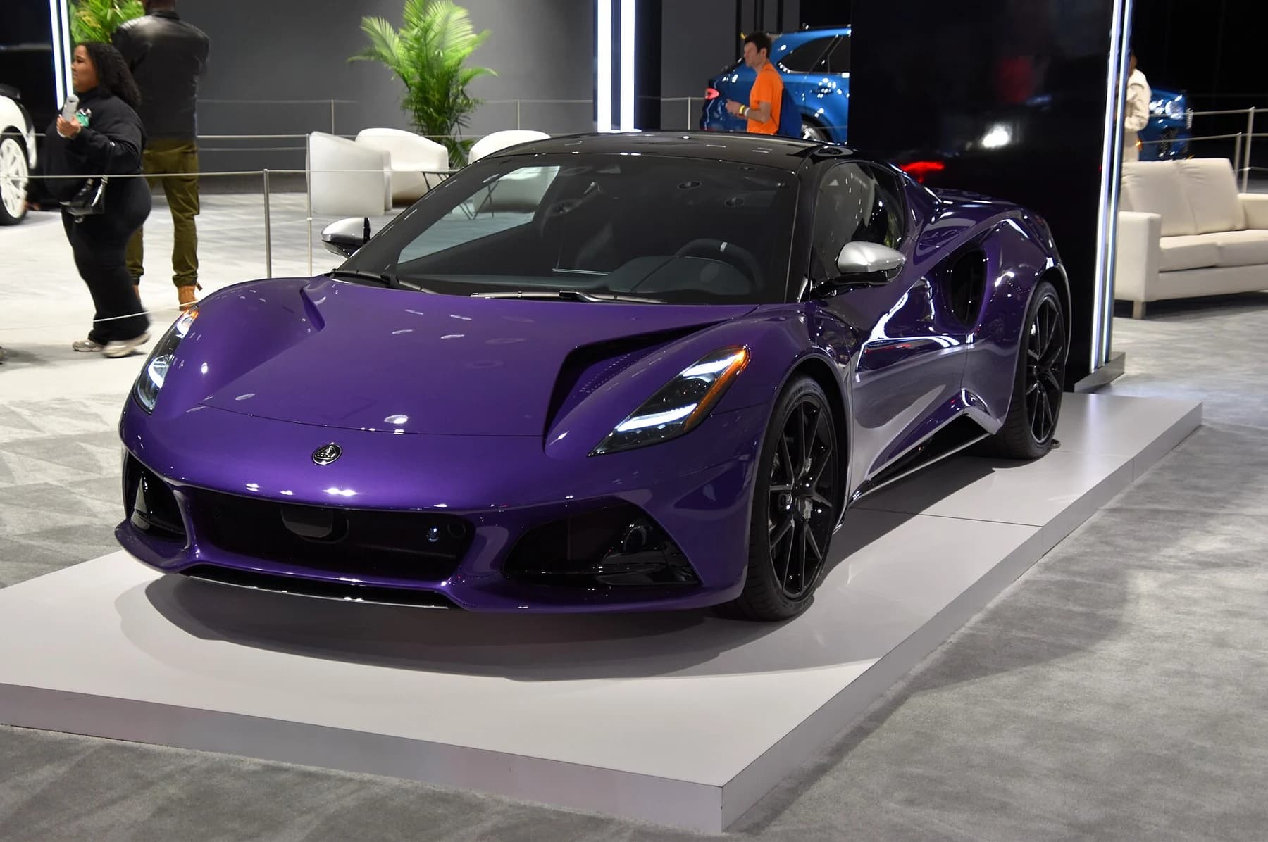 A purple Lotus Emira V6 SE seen from the front quarter on an indoor motor-show stand, with its low nose and black wheels visible.