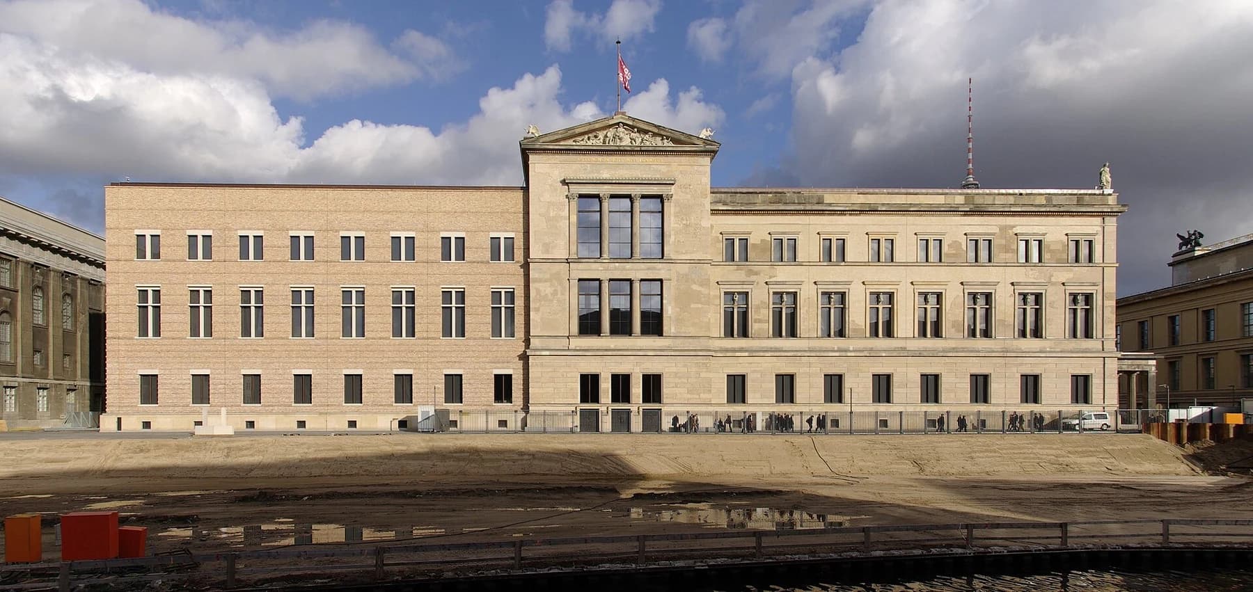 The long western facade of Berlin’s Neues Museum beneath sunlit clouds, with unfinished ground and works visible in the foreground.