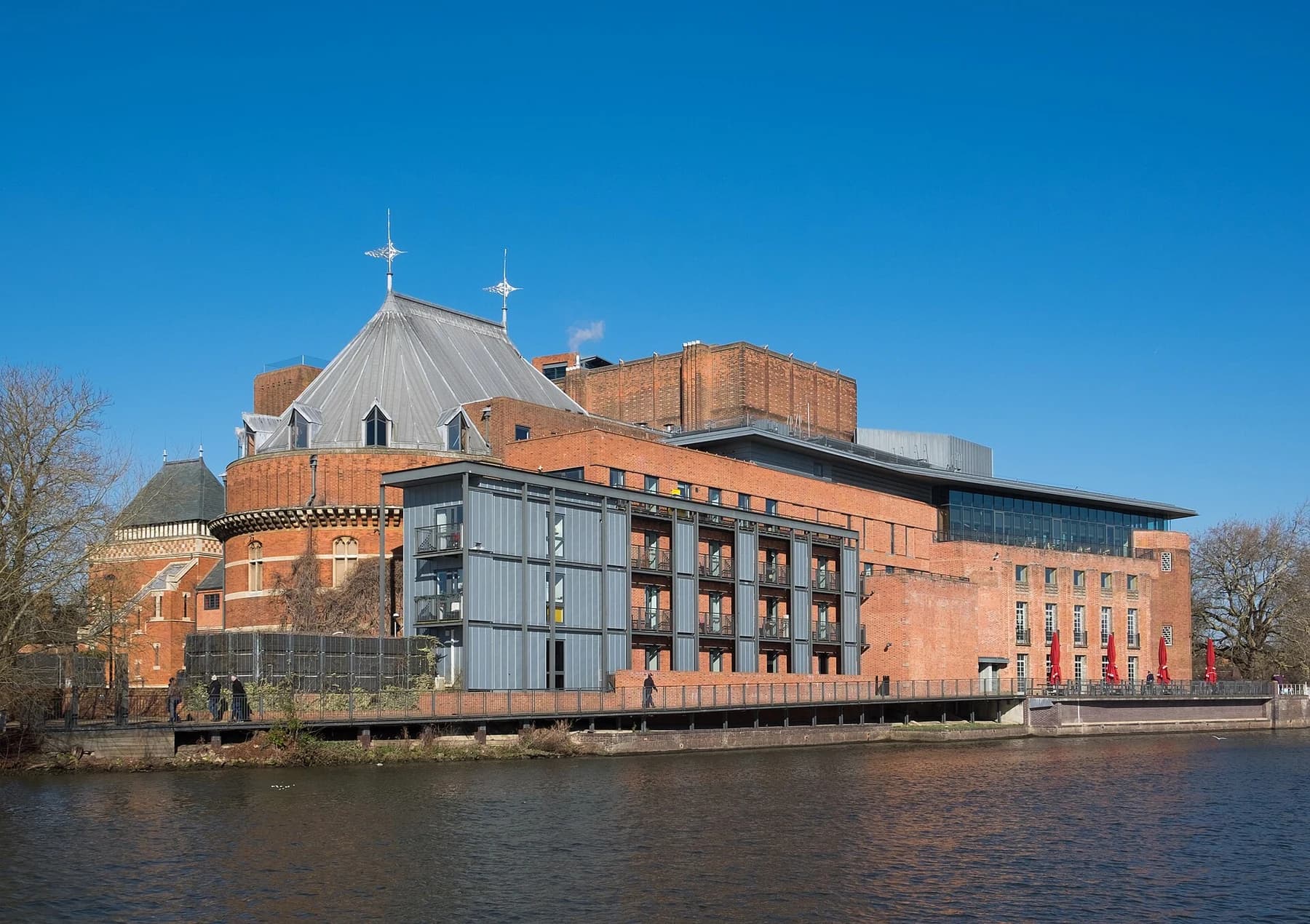 The Royal Shakespeare Theatre complex viewed across the River Avon from the south, with brick walls and the Swan Theatre visible under a clear sky.