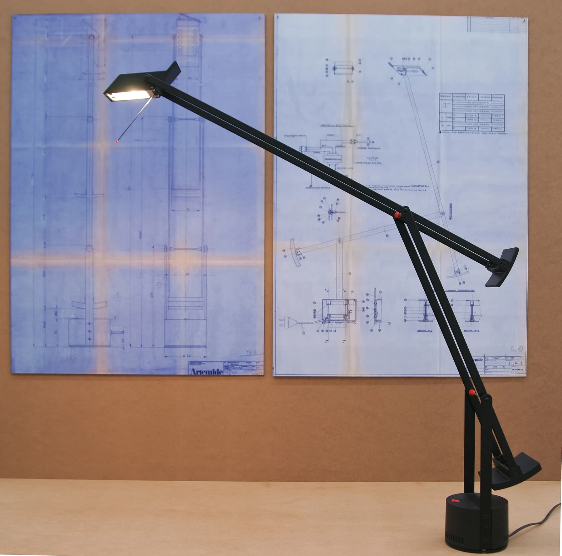 A black Tizio desk lamp with extended counterweighted arms, lit above a wooden desk with technical drawings pinned behind it.