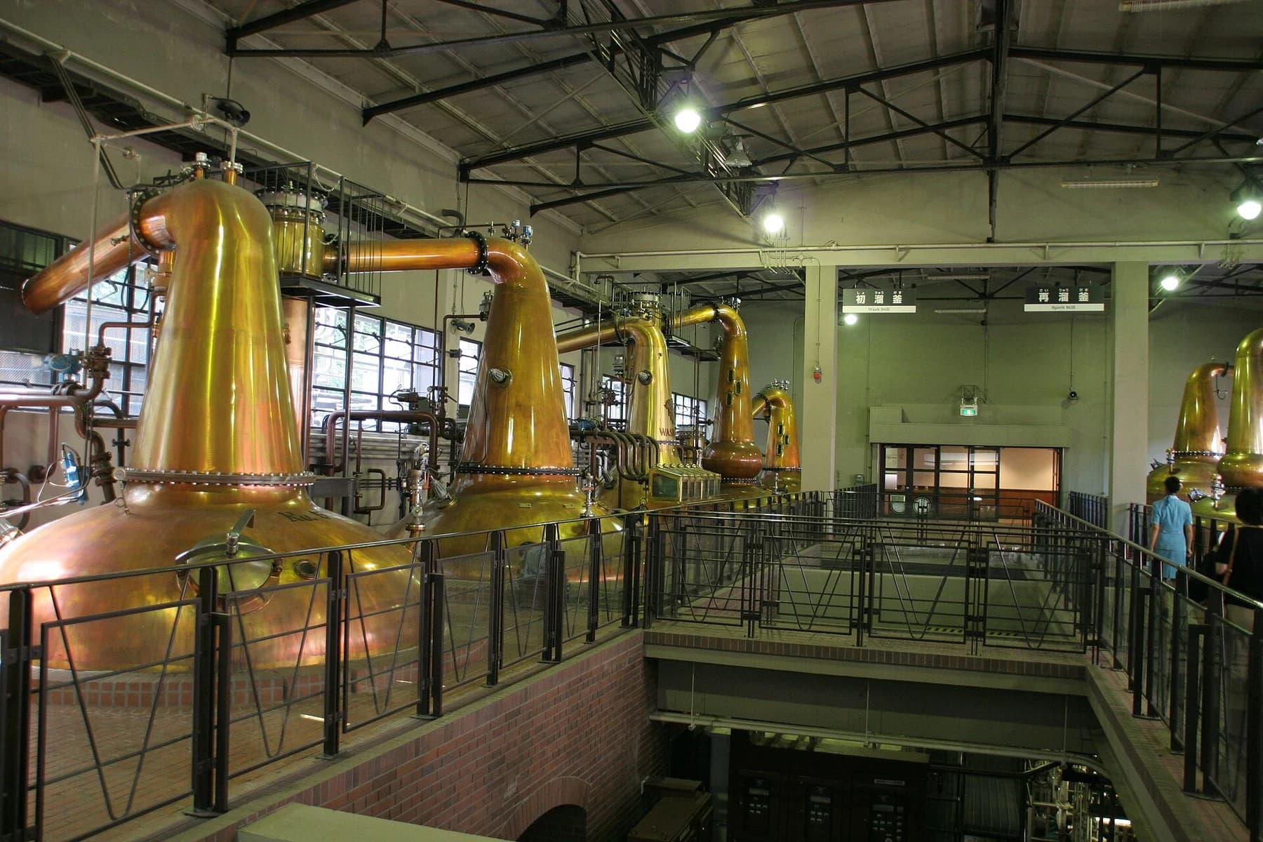 A row of copper pot stills inside Yamazaki distillery, seen beside a raised visitor walkway beneath the steel roof structure.