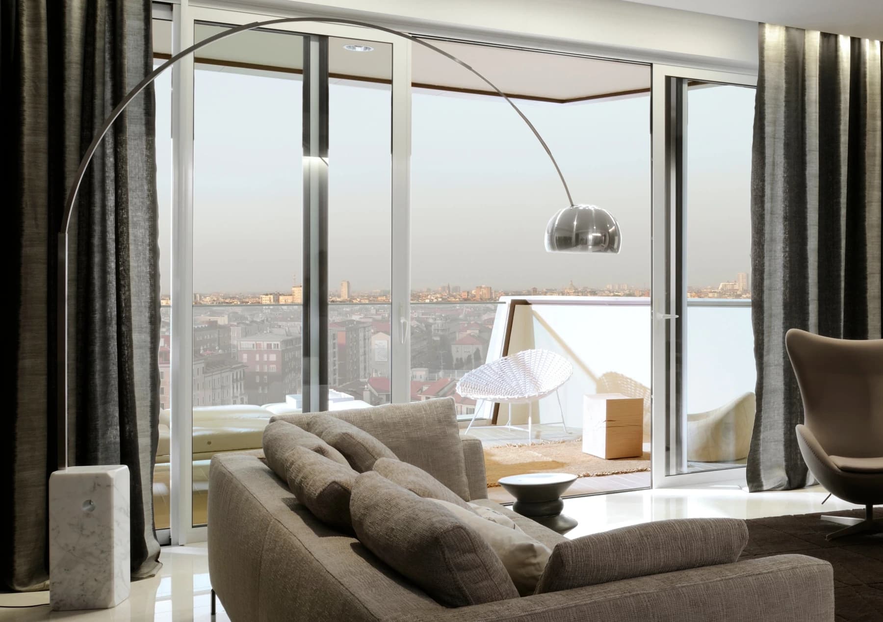 A Flos Arco floor lamp reaches across a contemporary living room from its marble base beside a sofa, with the full metal arch visible against large windows.