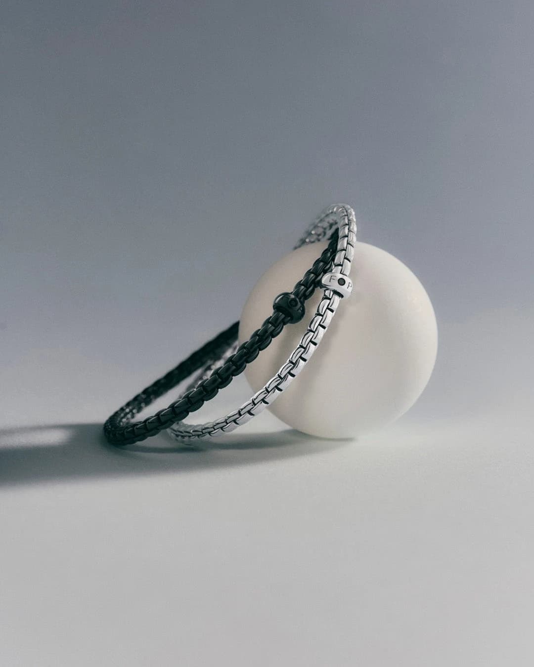 FOPE Eka Black and Eka flexible gold bracelets arranged together on a white cylindrical display, showing the contrast between their dark and light metal surfaces.