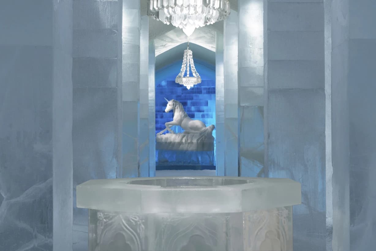 The 2014 ICEHOTEL main hall with translucent ice columns, hanging ice chandeliers and a horse sculpture framed by blue-lit openings in the distance.