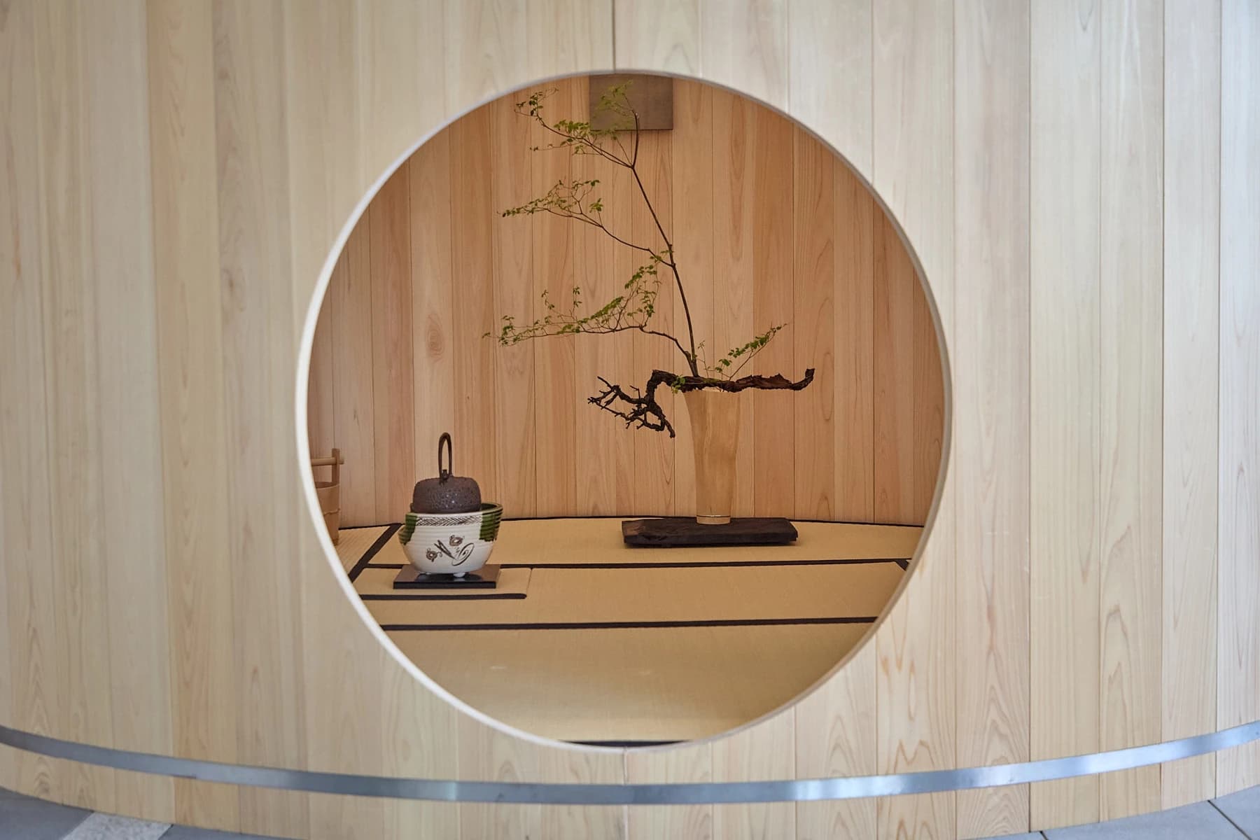 Shuji Nakagawa’s cylindrical wooden tea room with its circular doorway, curved timber walls and external hoop visible, opening onto a small interior with a mat and flowers.