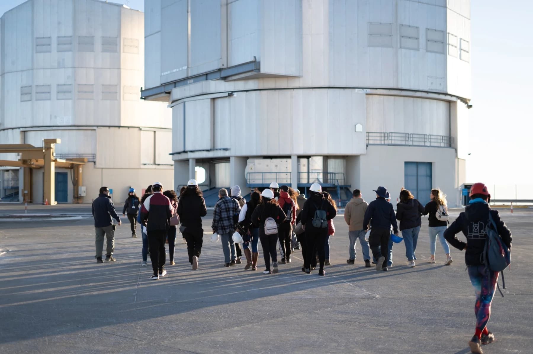 Visitors walking across the Paranal Observatory platform beside the large white Very Large Telescope enclosures in the dry Chilean landscape during a daytime visit.