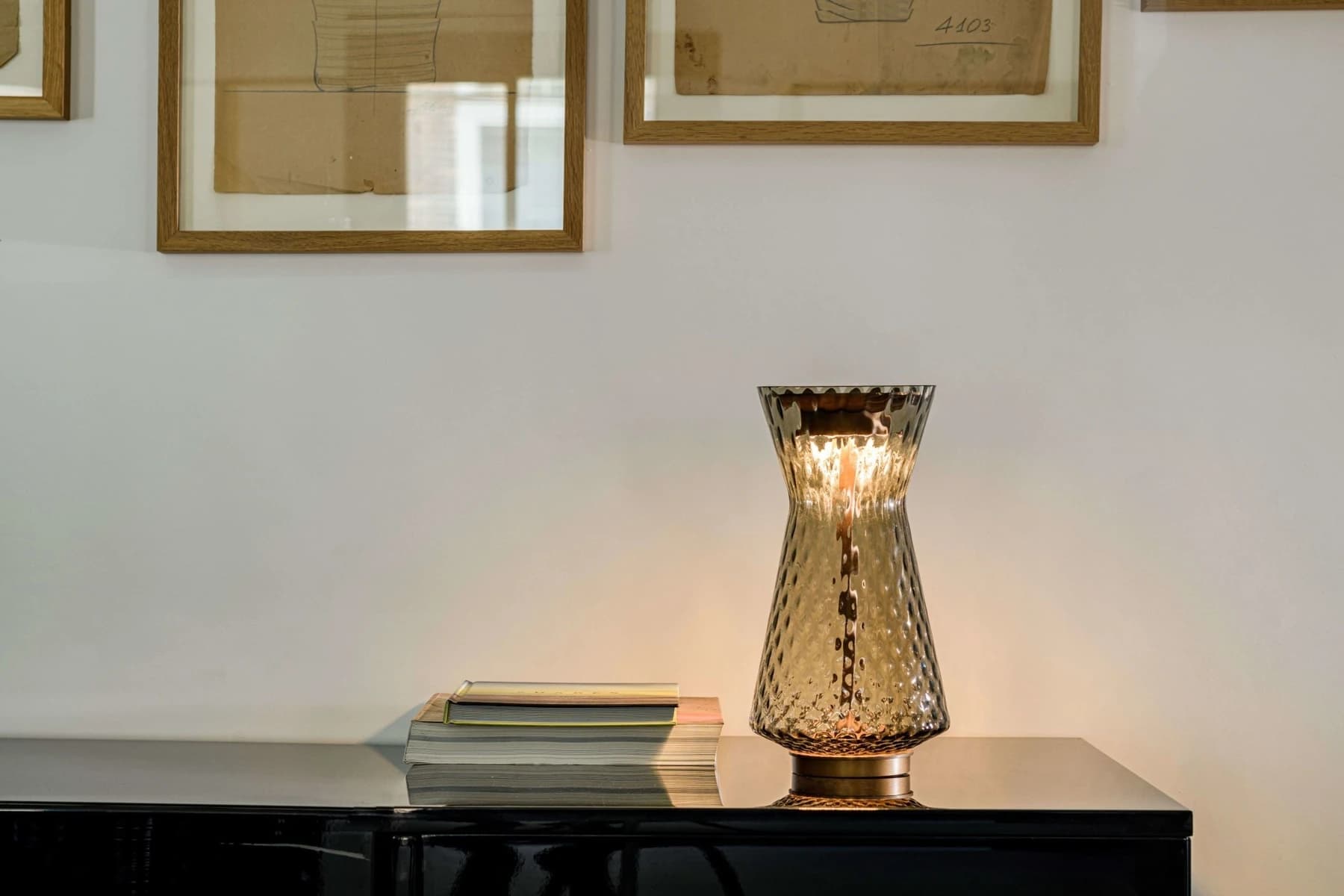 An amber Venini Tiara Luce table lamp glowing on a dark sideboard beneath framed artwork, with its rounded quilted glass body and warm reflected light visible.