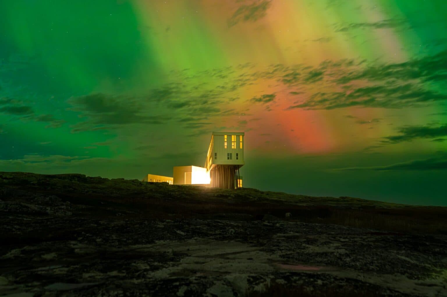 Fogo Island Inn illuminated at night beneath green and orange northern lights above a dark rocky landscape.