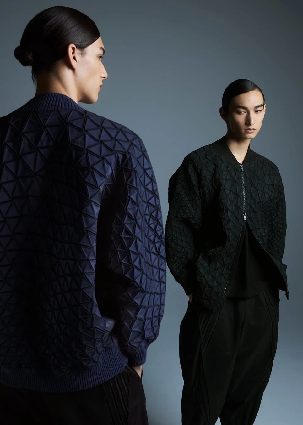 Two models wearing dark sculptural TYPE-V garments, with the geometric three-dimensional textile visible across the back of a blue jacket in the foreground.
