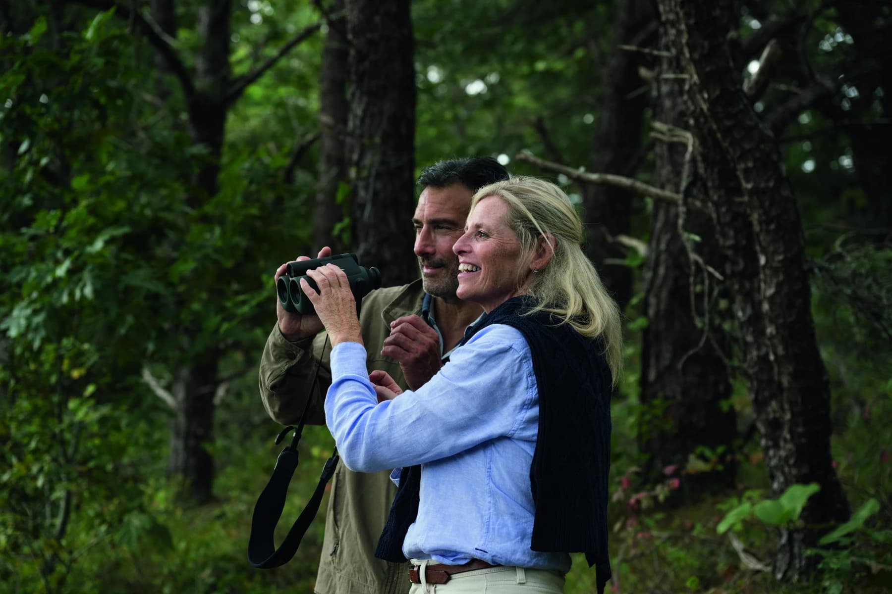 Two people in a green wooded landscape holding Swarovski Optik AX Visio binoculars, in the manufacturer’s supplied outdoor press photograph.