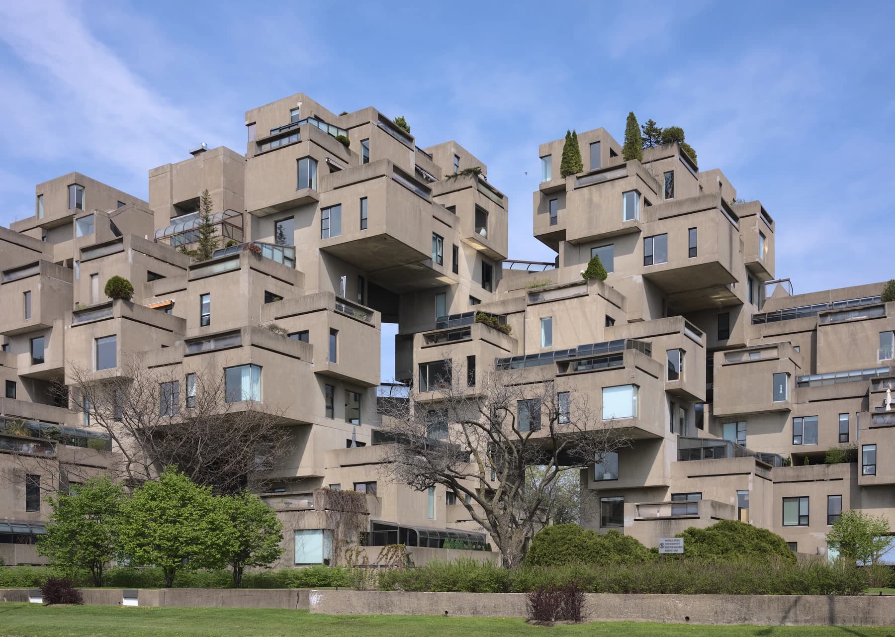 Habitat 67 in Montréal viewed from ground level, showing stacked concrete homes, balconies, planted terraces and projecting modules beneath a blue and lavender sky.