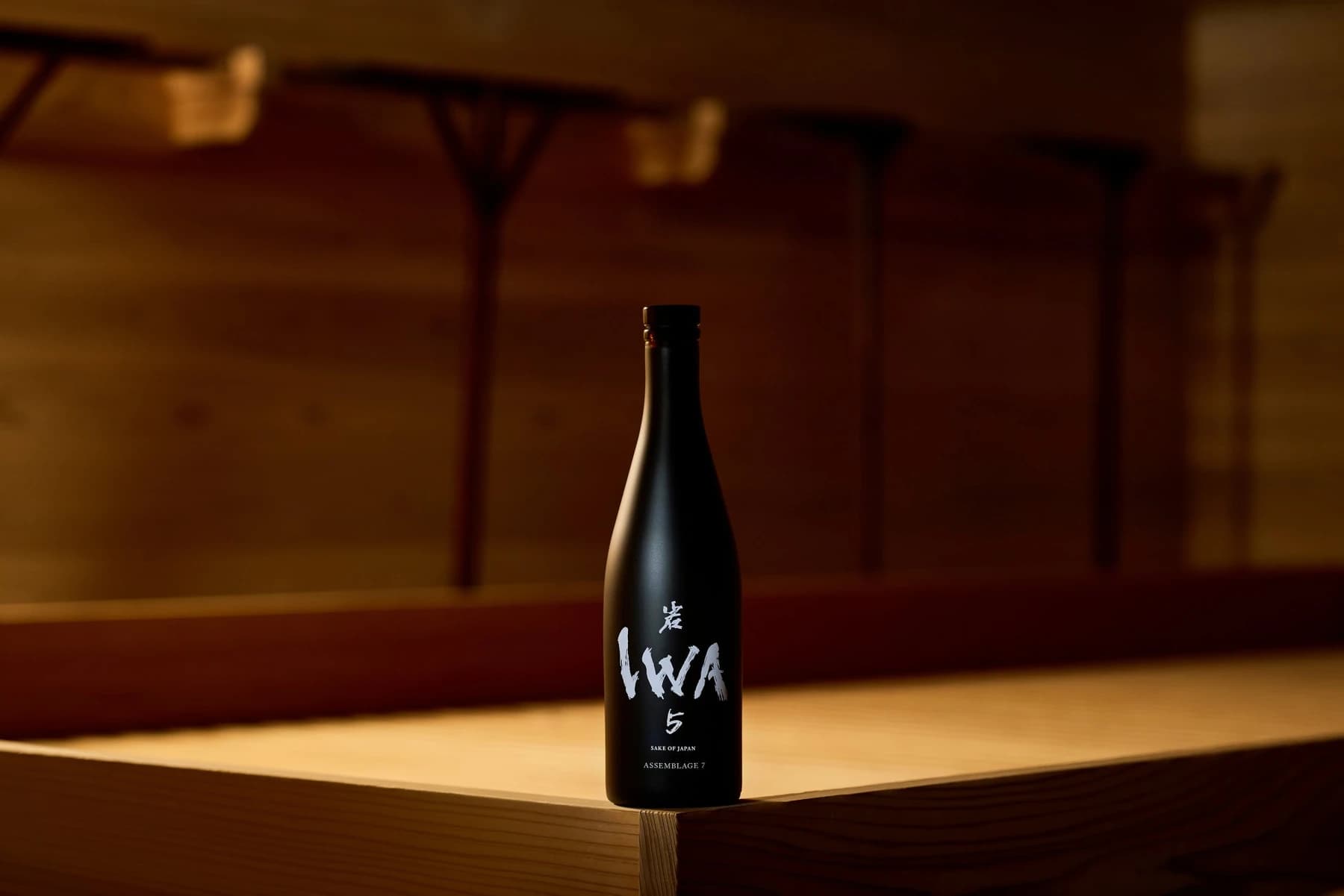 A dark IWA 5 sake bottle standing on a warmly lit wooden surface, with a small assemblage number beneath its white calligraphic mark.