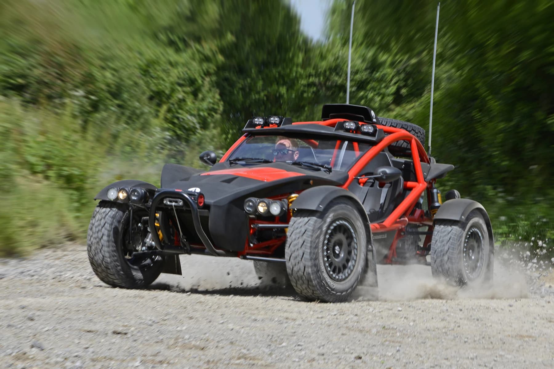 A red-framed Ariel Nomad 2 driving across a loose-surface track with a helmeted occupant, exposed suspension and dust behind the rear wheels.