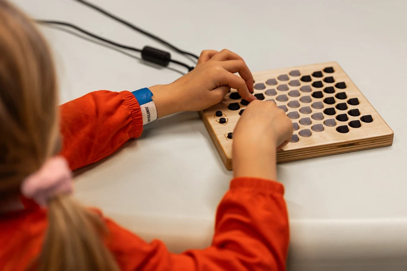 A participant in an orange-red blouse playing a wooden electronic instrument with rows of round buttons at SoundLAB, with hands and the physical controls in focus.